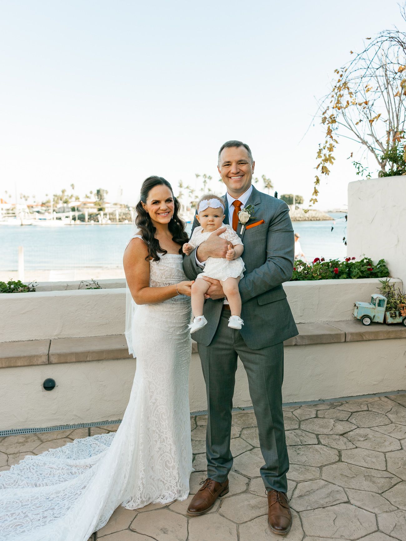 Family in formal attire poses together on waterfront terrace during wedding celebration.