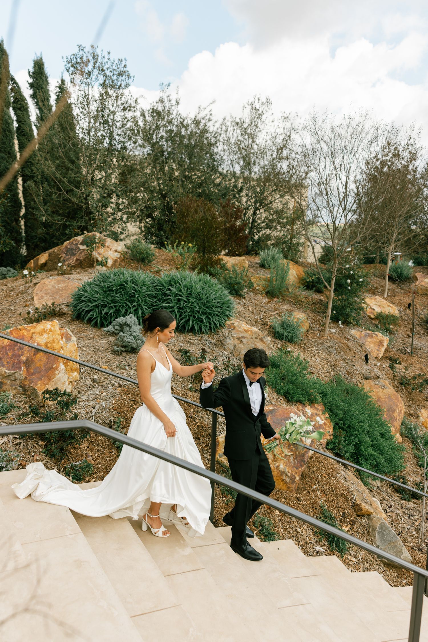 A couple descends stone steps at an outdoor venue surrounded by lush landscaping and greenery.