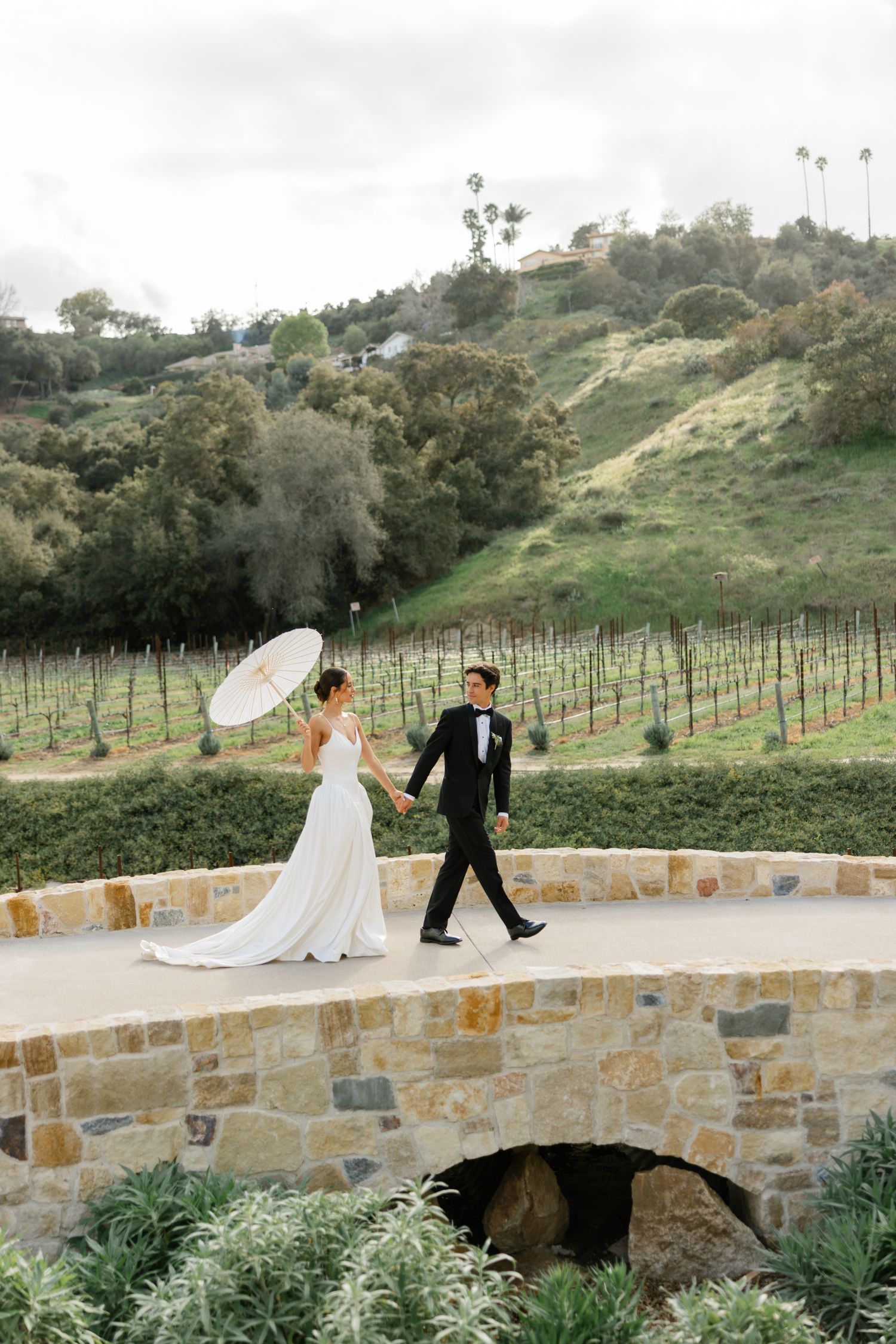A bride and groom walking along a stone wall pathway with vineyard and rolling hills in the background.