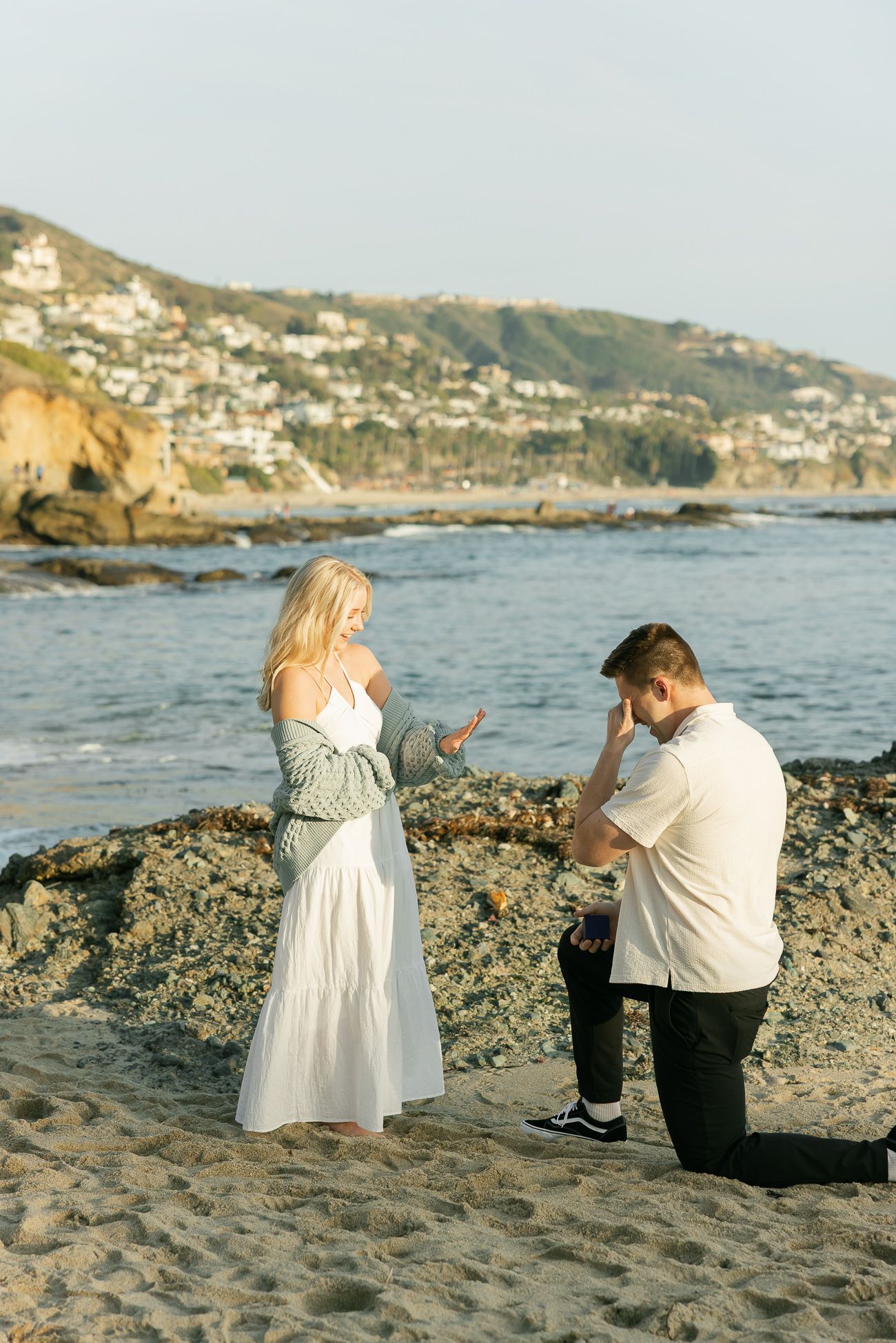 A surprise proposal moment unfolds on a scenic beach with coastal homes visible in the background.