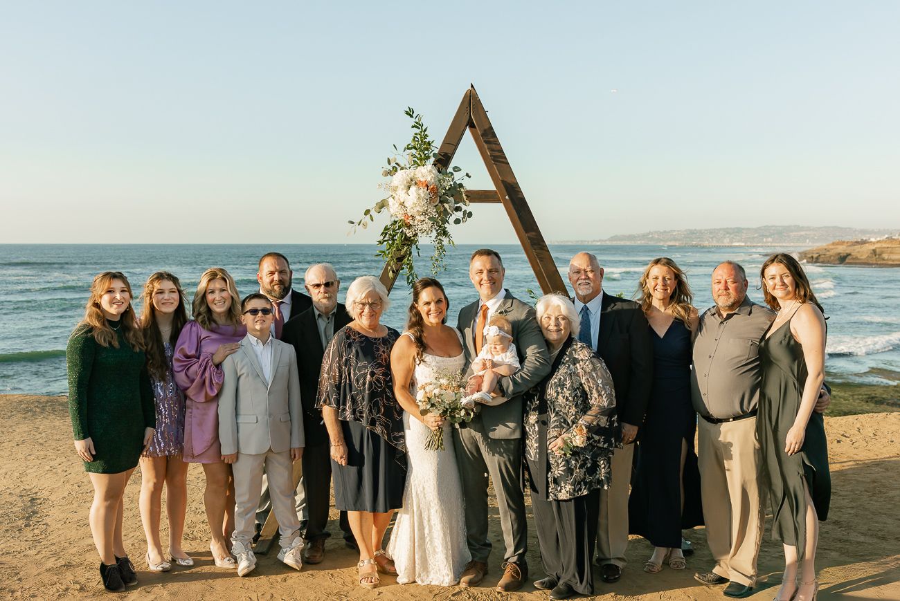 Wedding party gathered under wooden triangular arch on coastal cliffside during sunset ceremony.