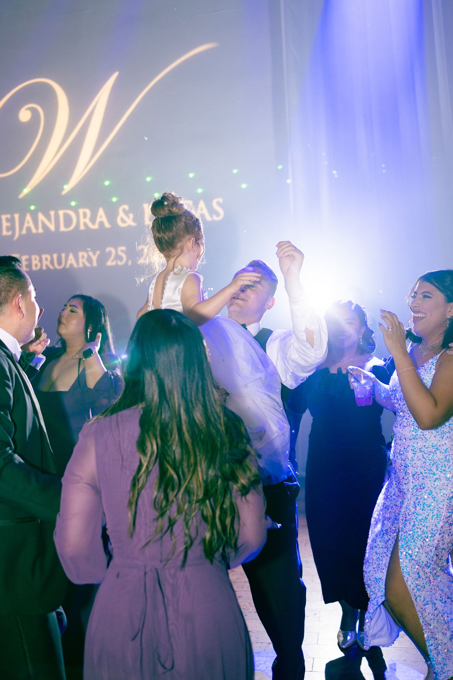 Wedding guests dancing and celebrating at a reception with dramatic blue lighting and monogram display.