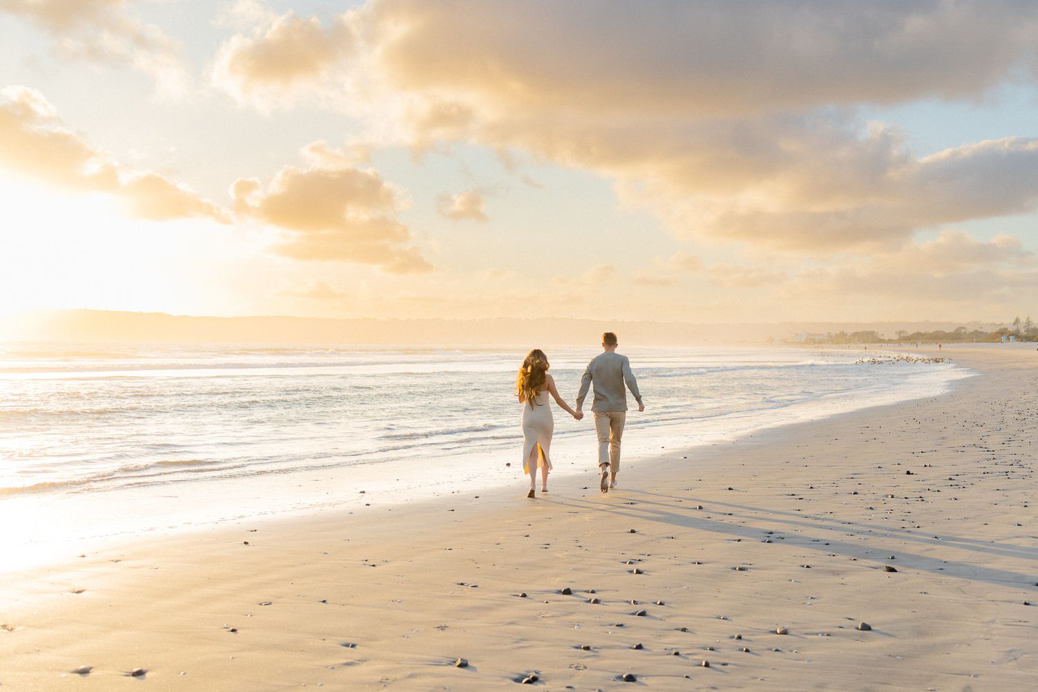A couple walks hand in hand on a sandy beach at sunset with golden clouds reflecting on the wet sand.