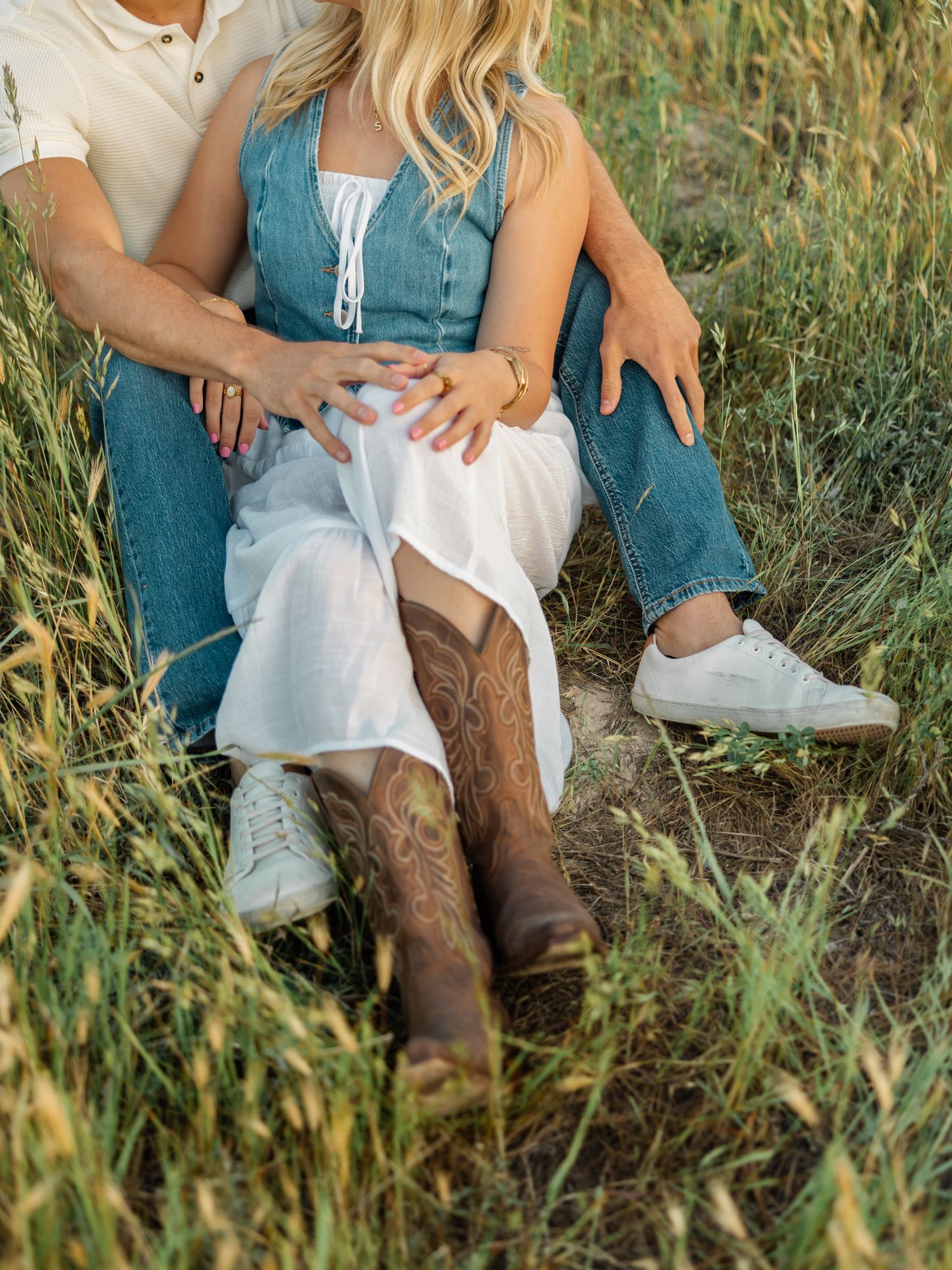 Couple sitting together in tall grass wearing jeans and boots showing romantic country style engagement poses.
