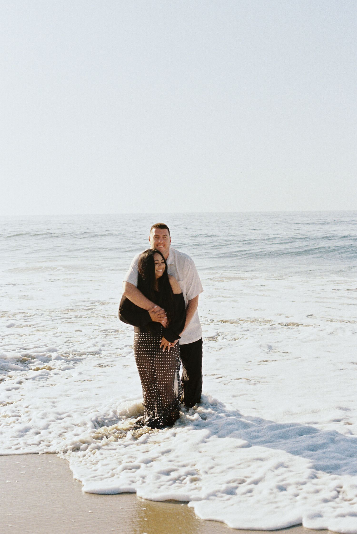 A couple embraces while standing in the ocean waves on a sunny beach.