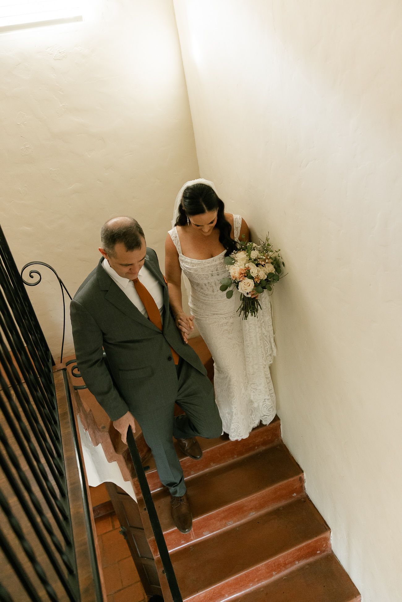 A couple descends a staircase together, with the bride in white and groom in gray suit holding hands.