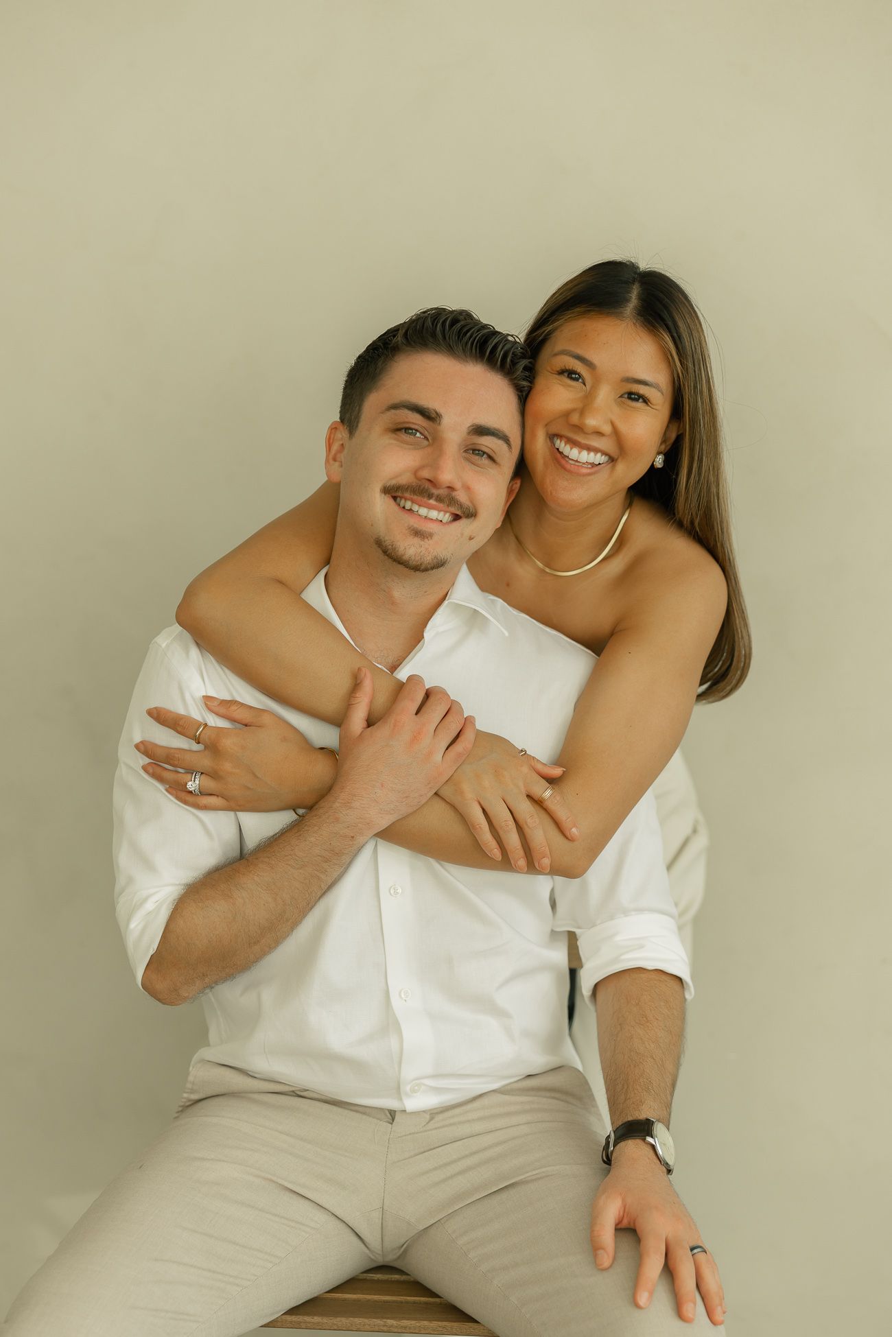 A couple wearing white embraces and smiles while posing against a beige wall in a romantic portrait.