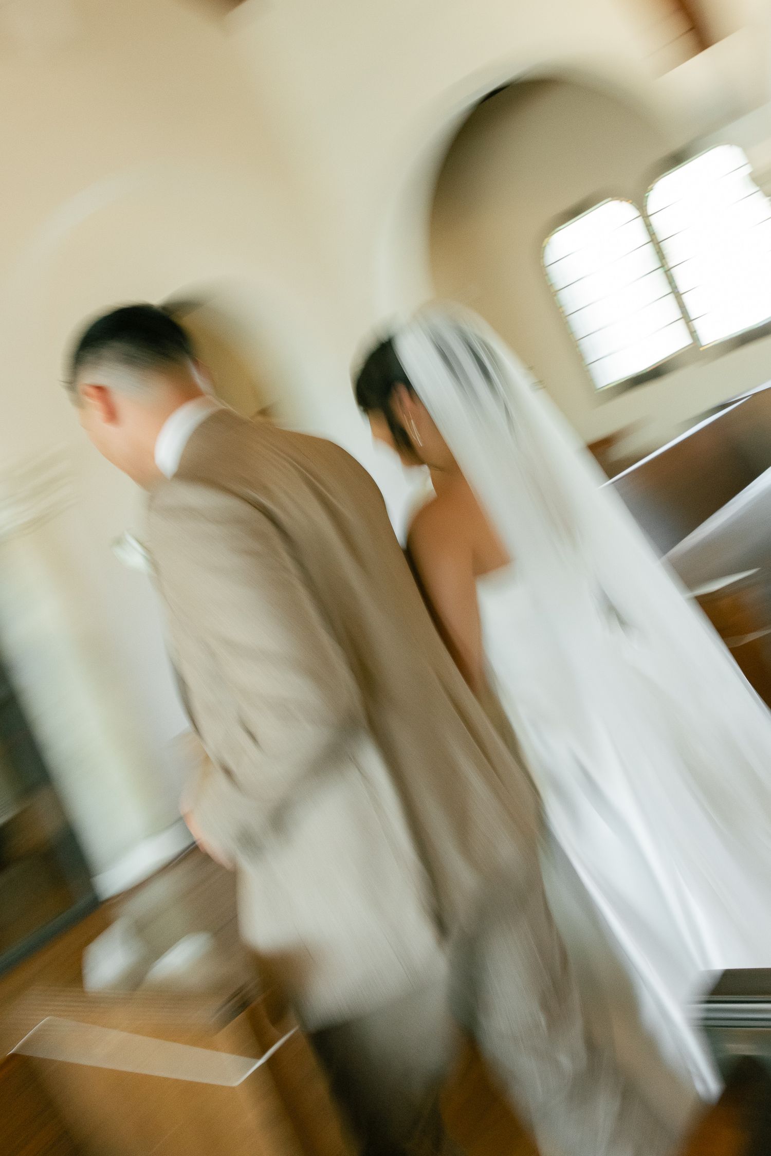 Motion-blurred image shows a bride in white veil and groom in beige suit walking through an interior space.