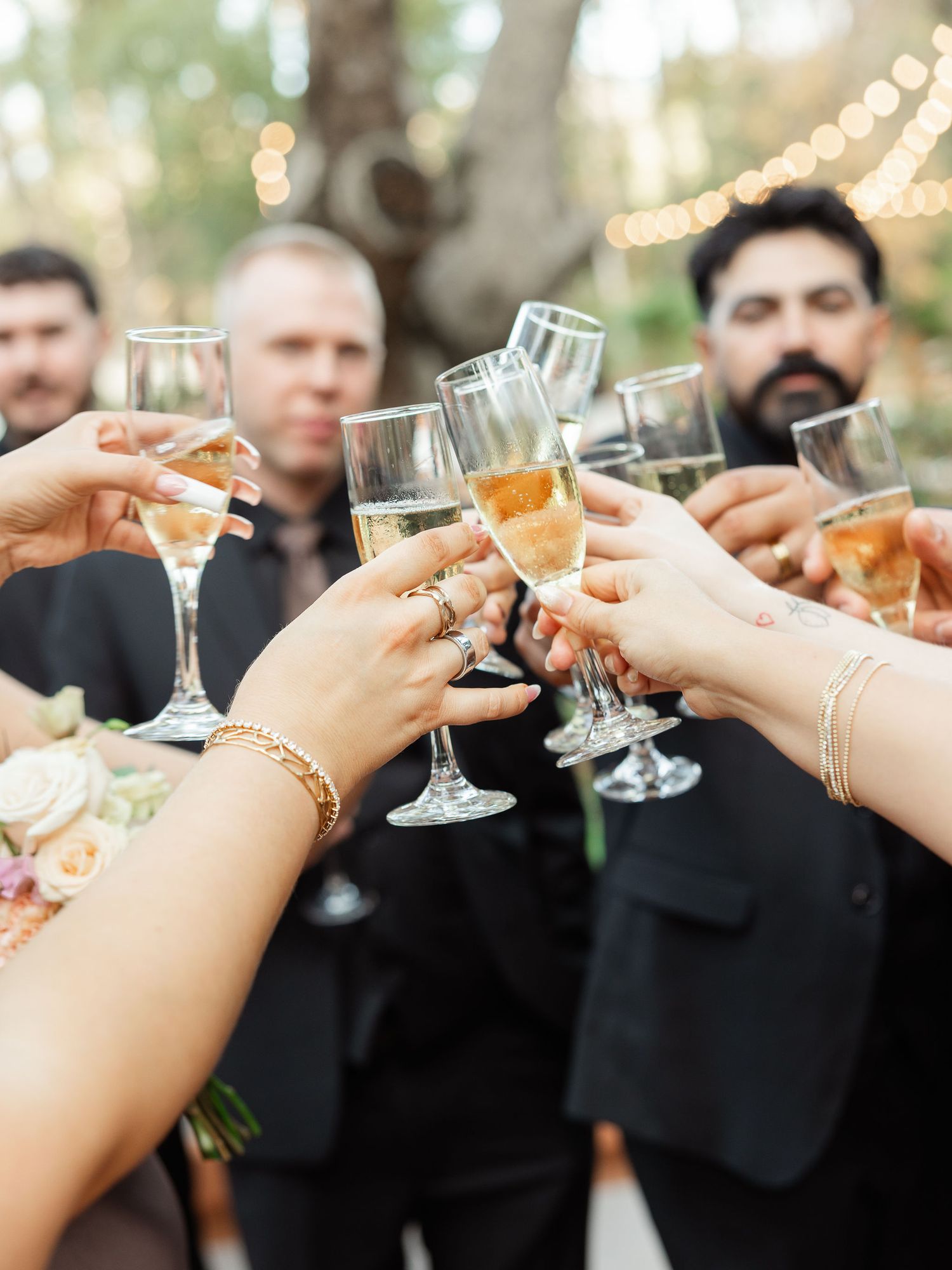 Wedding guests raise champagne glasses in a celebratory toast during an outdoor reception with twinkling lights.