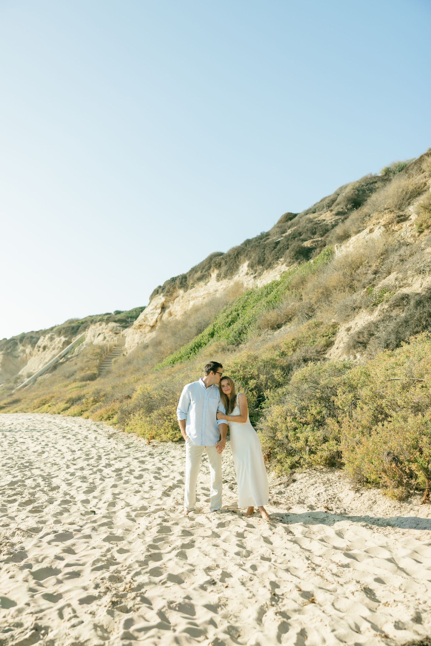 A couple in wedding attire walks along a sandy beach path with cliffs in the background during golden hour.