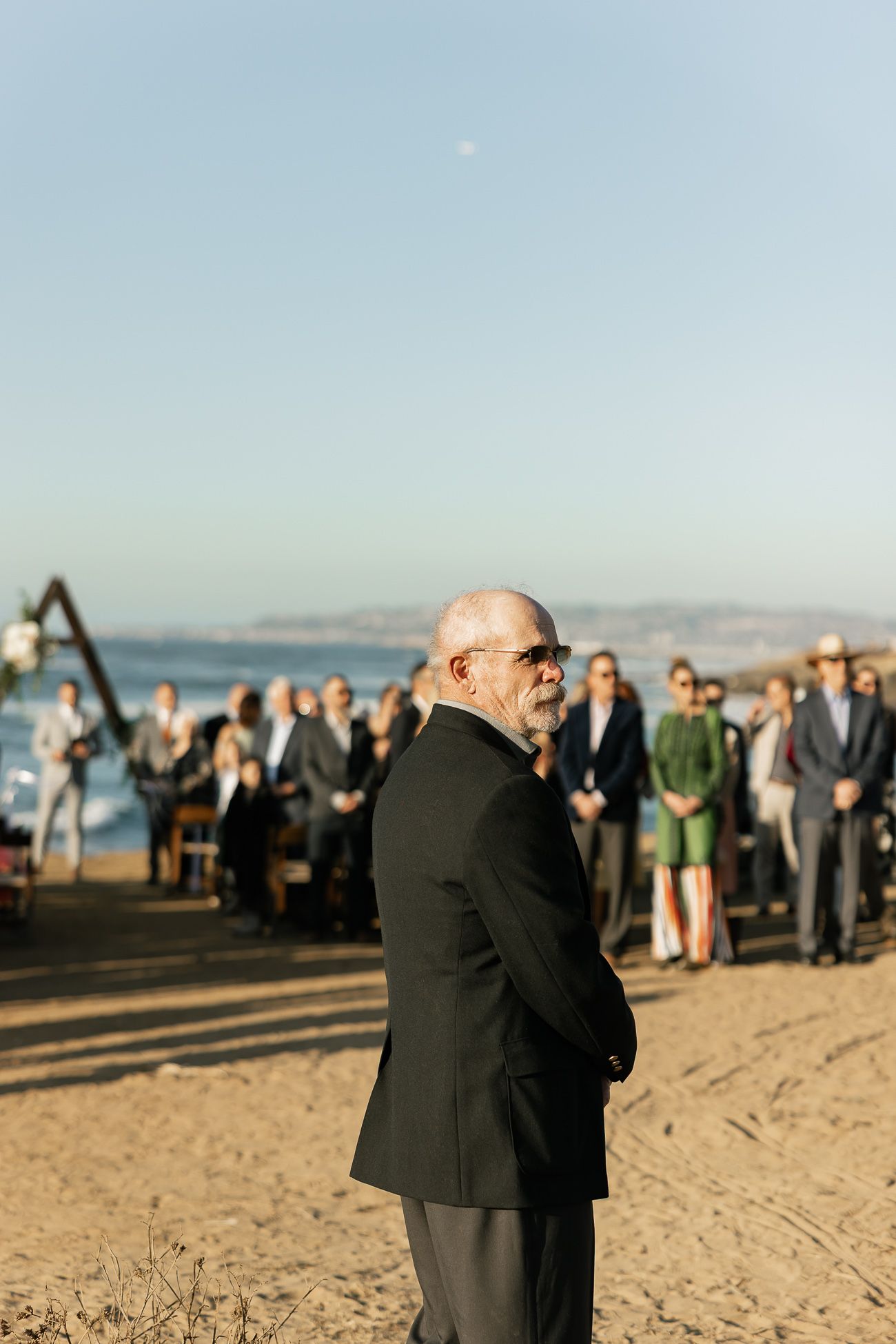 A wedding ceremony on a sunny beach with guests gathered and someone standing in formal attire by the shore.