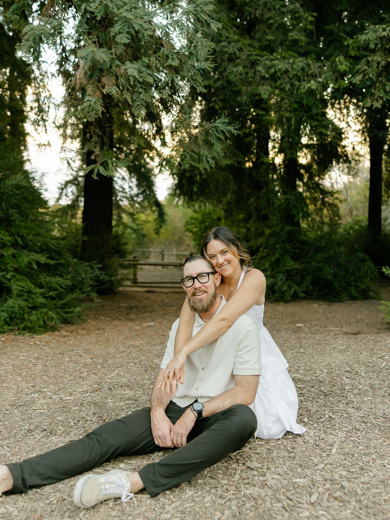 A couple sits together on a dirt path surrounded by evergreen trees in a natural outdoor setting at sunset.