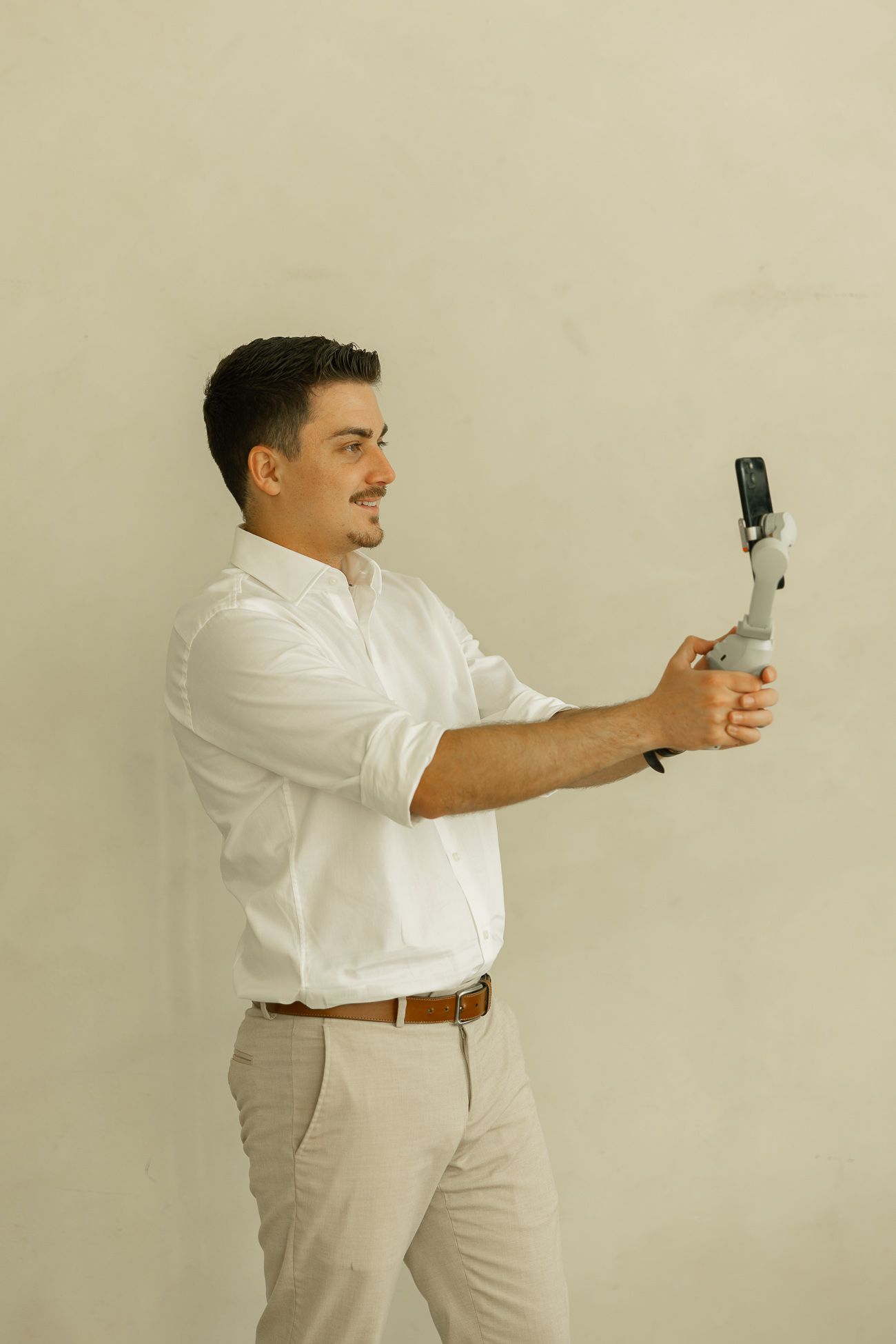 Professional in white shirt and khakis stands sideways while holding a phone gimbal stabilizer against beige wall.