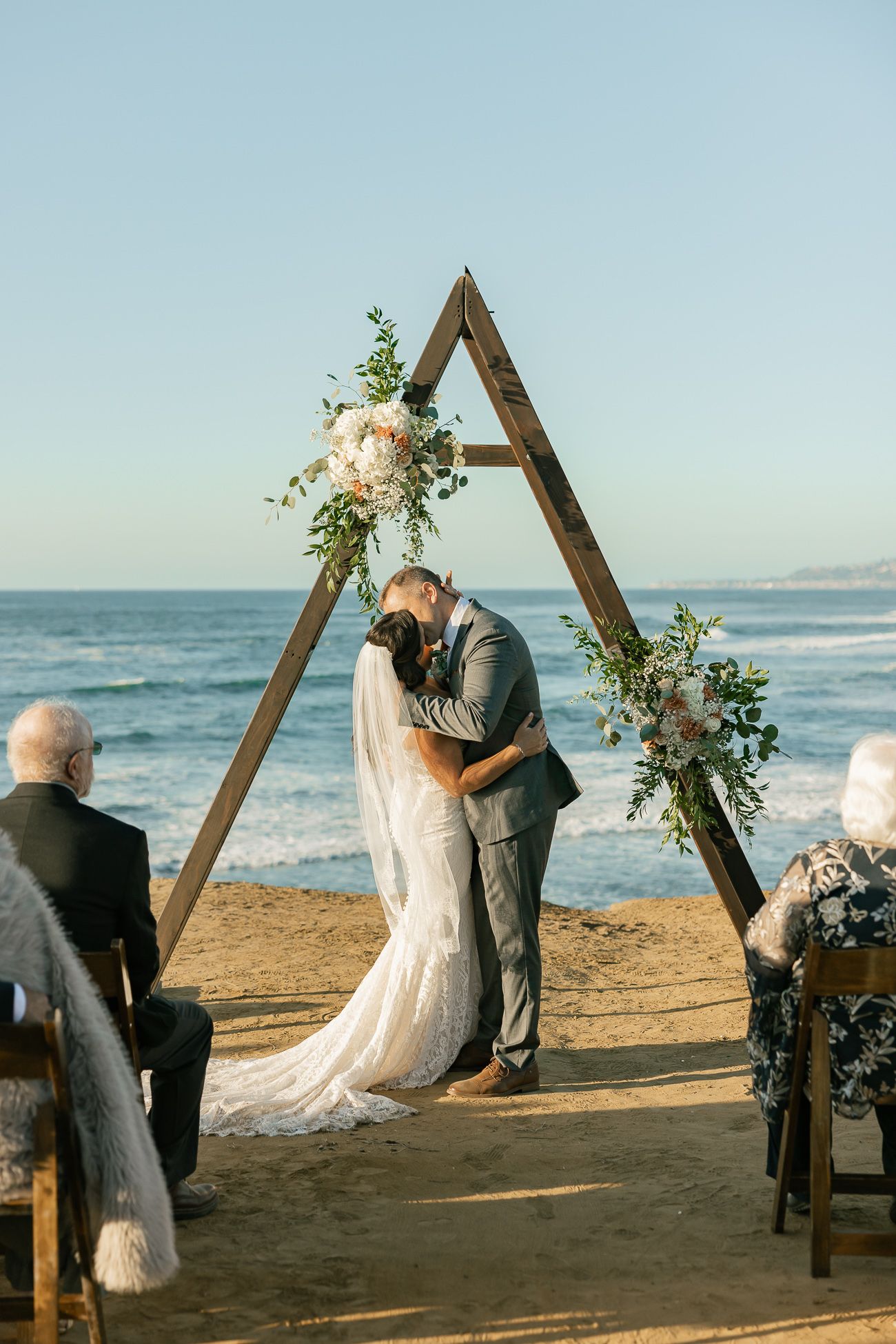 Romantic beach wedding ceremony under wooden triangle arch decorated with white flowers and greenery.