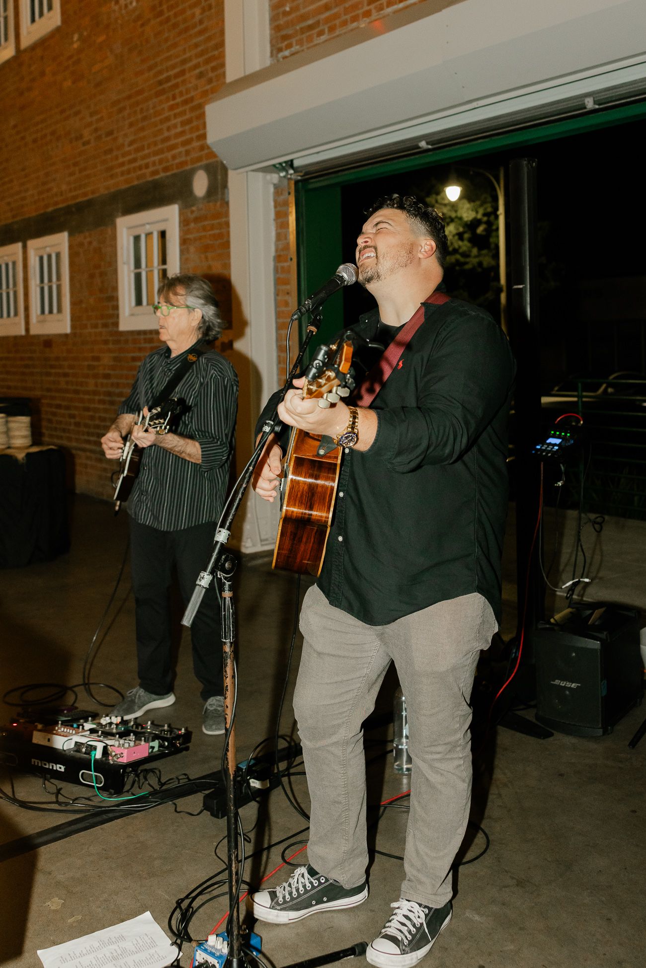 Musicians performing with guitars at a nighttime event against a brick building exterior.