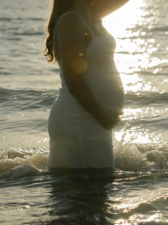 Silhouette of pregnant woman's belly in water at sunset with light sparkling on gentle waves.