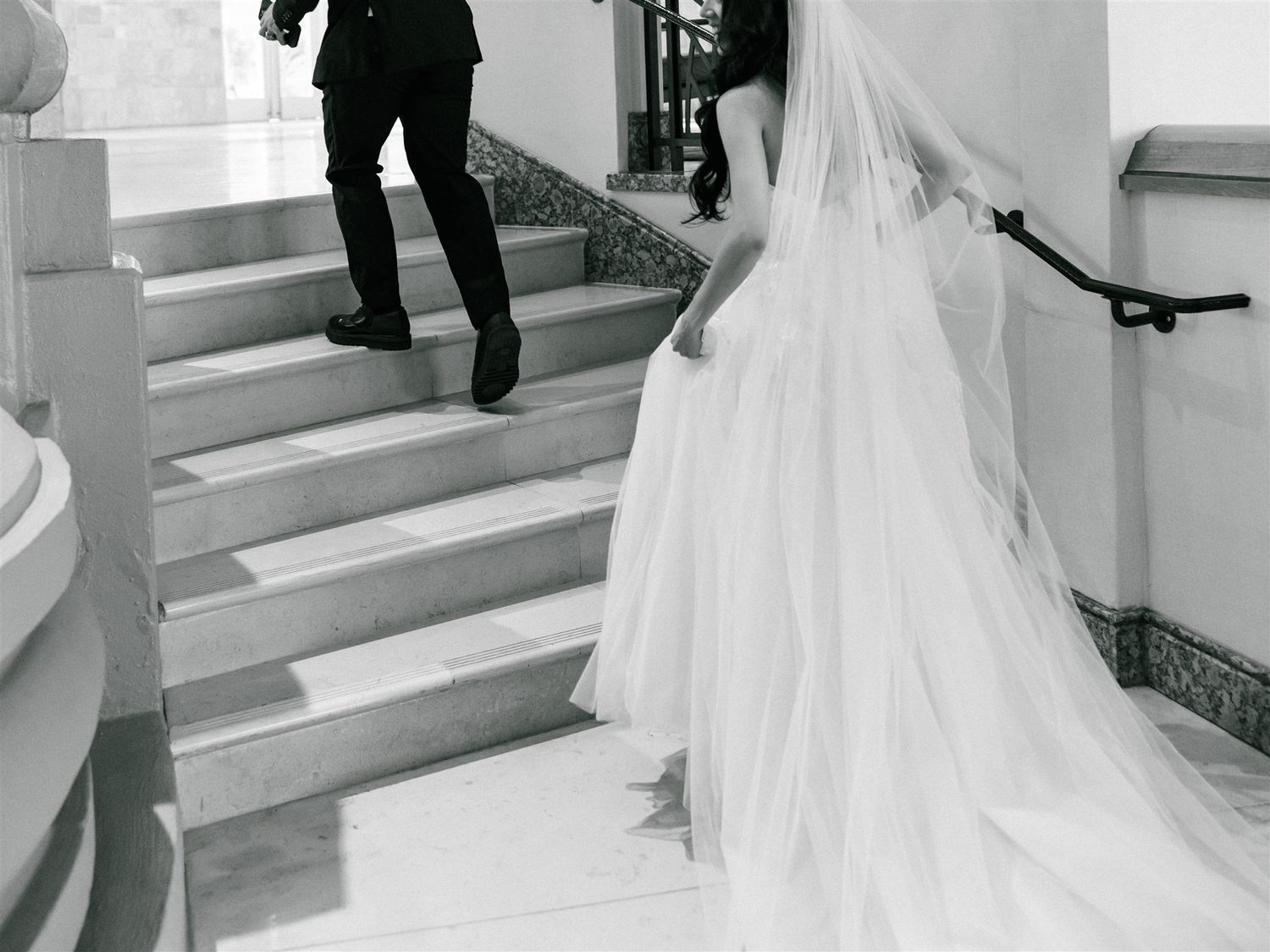 A flowing wedding dress train follows up marble stairs in black and white.