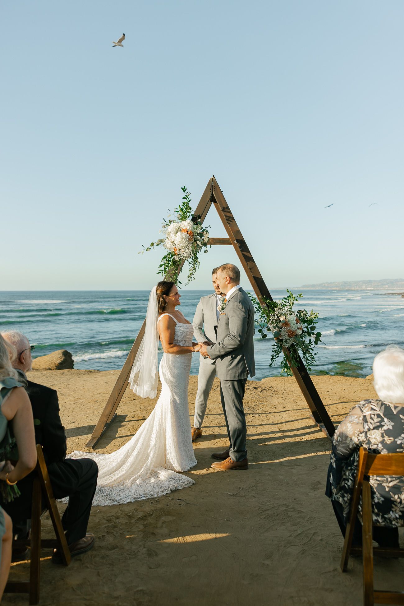 A wedding ceremony takes place under a wooden triangle arch on a beach at sunset with ocean waves in the background.