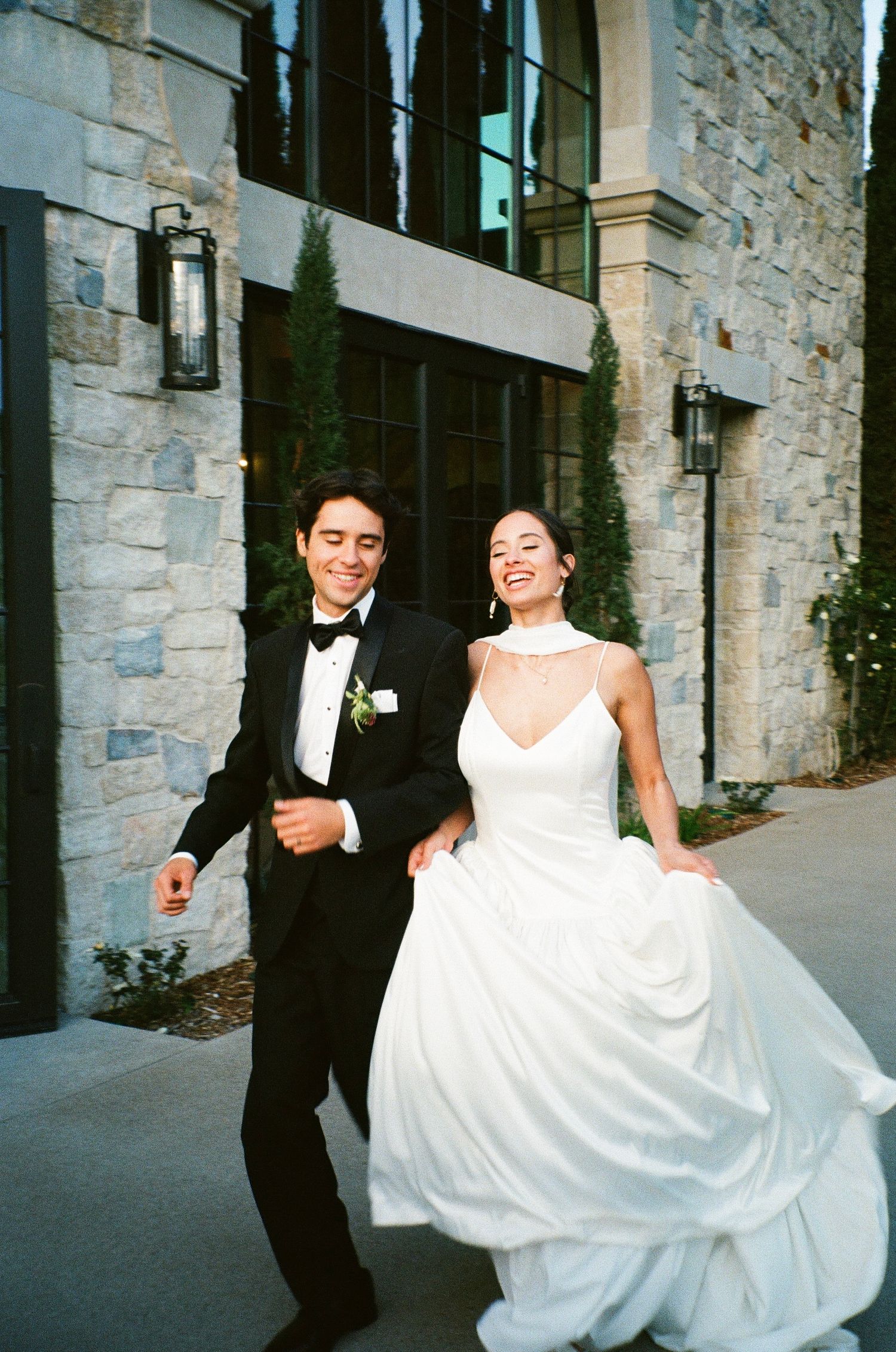 A joyful couple walking outside a stone building on their wedding day, with the bride in white and groom in a tuxedo.