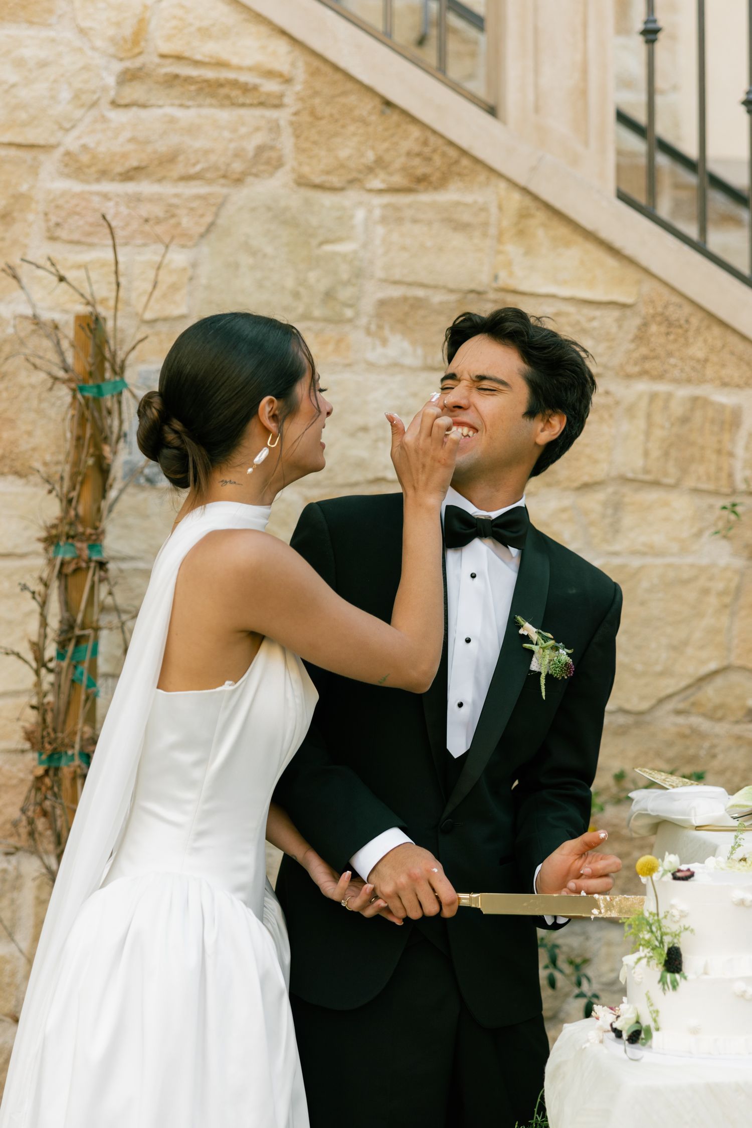 A newlywed couple smiles and shares wedding cake during their joyful reception outdoors against a stone wall.