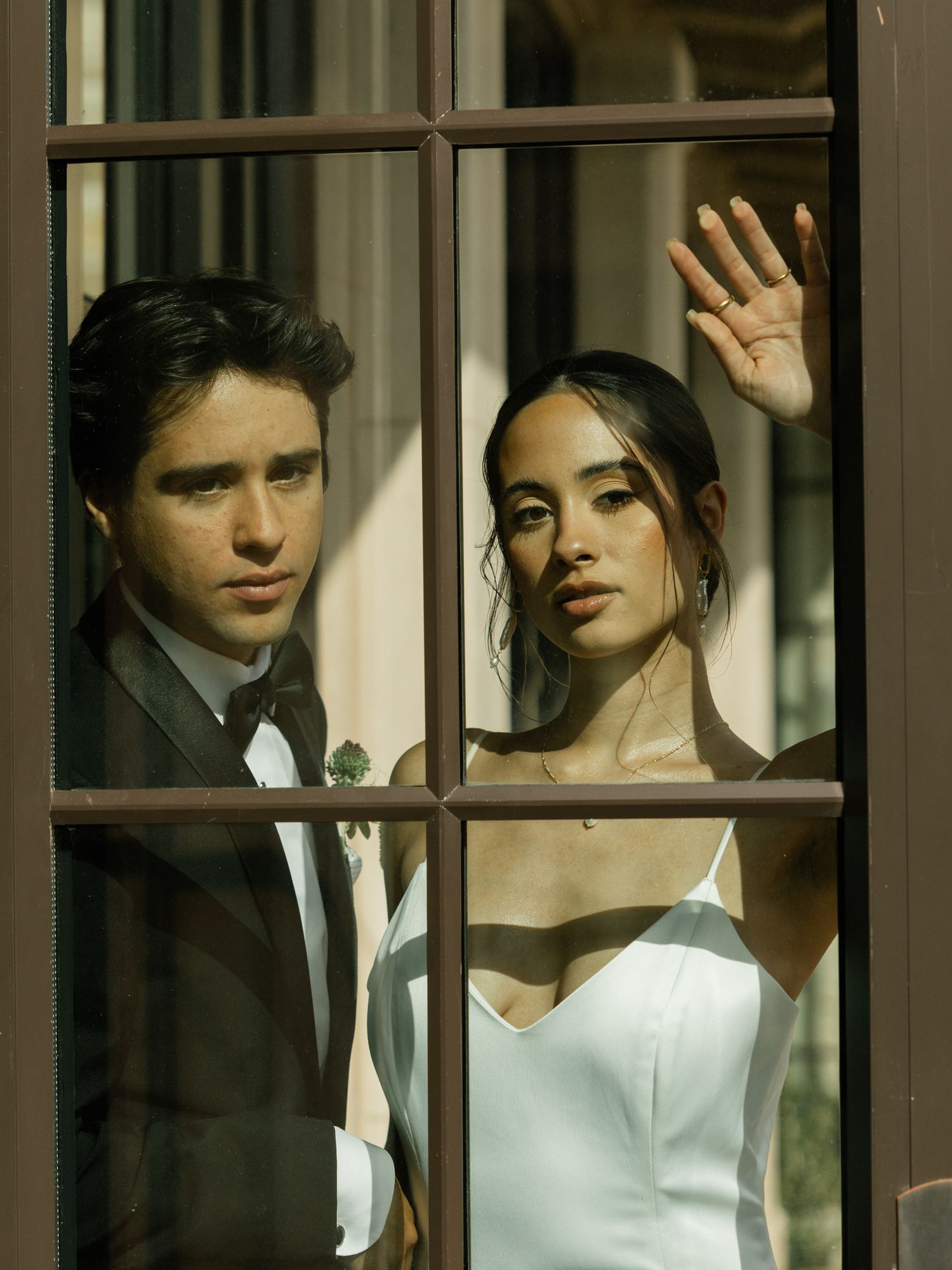 A romantic wedding portrait through a windowpane shows a couple posing in formal attire with warm lighting.