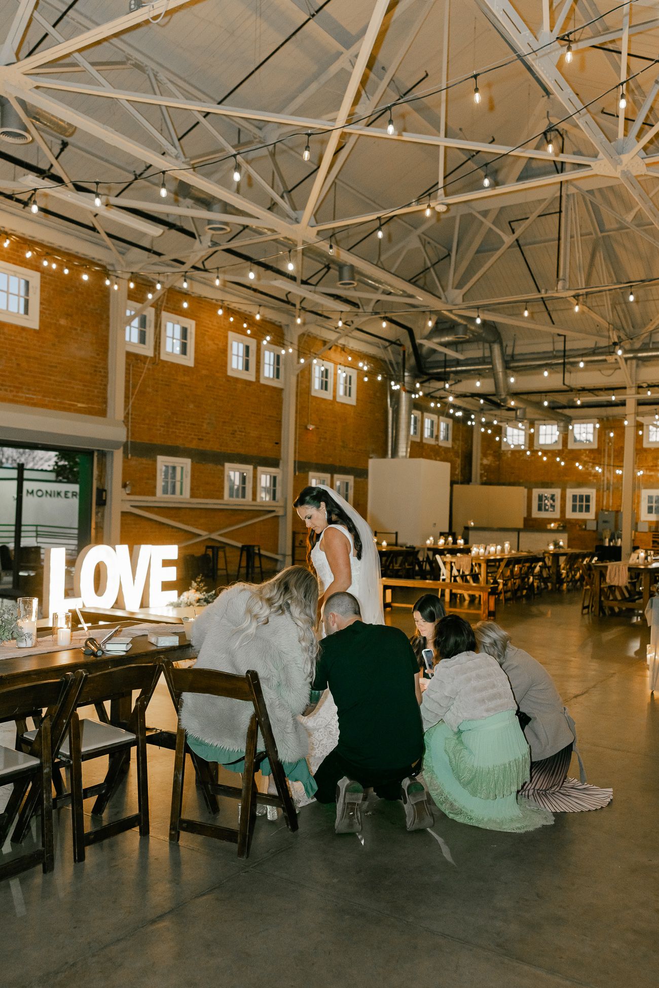 Wedding guests huddle around a table in an industrial venue with string lights and a glowing LOVE sign.