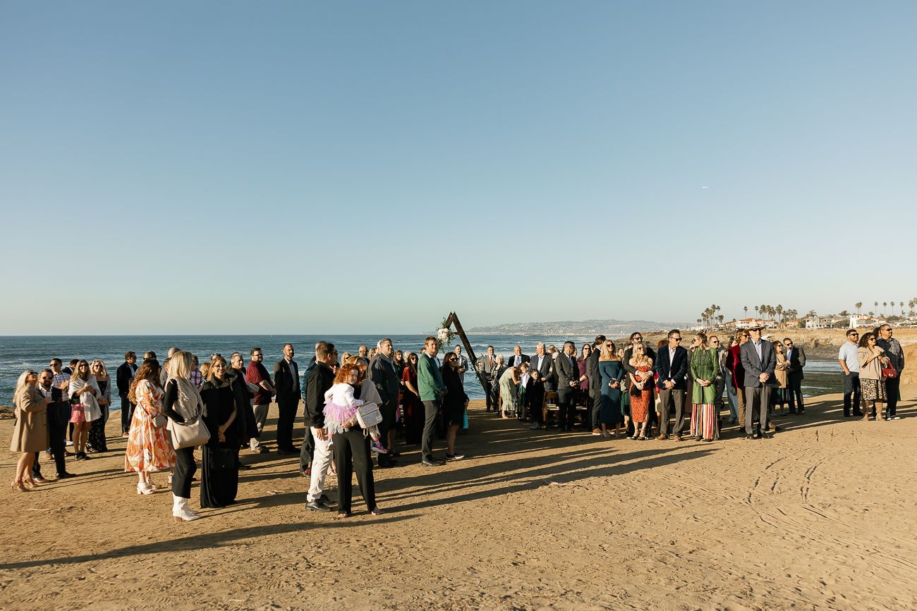 Wedding guests gathered on a sunny beach with ocean views and blue skies during an outdoor ceremony.