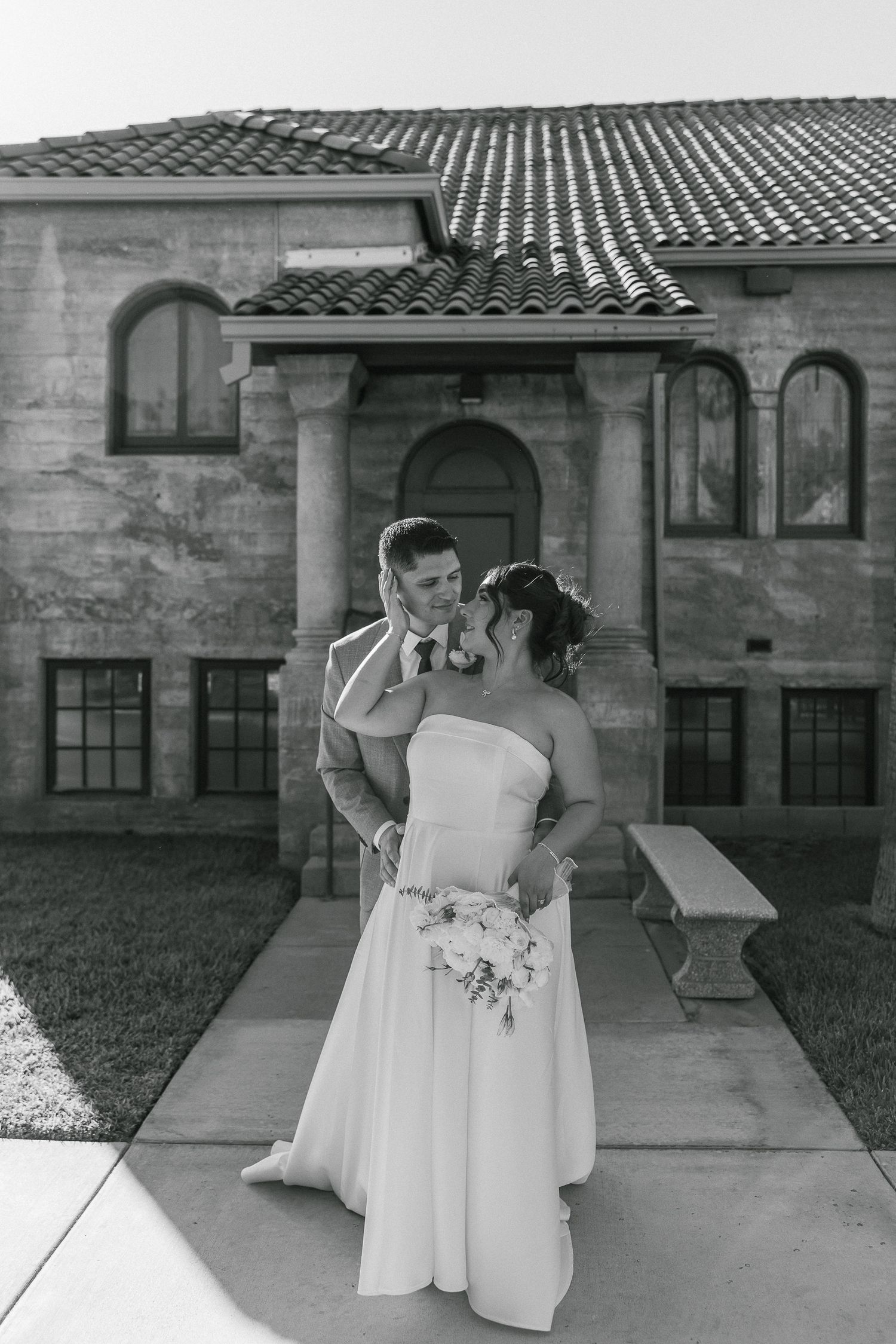 A couple in wedding attire stands together on a patio with Spanish-style architecture in black and white.