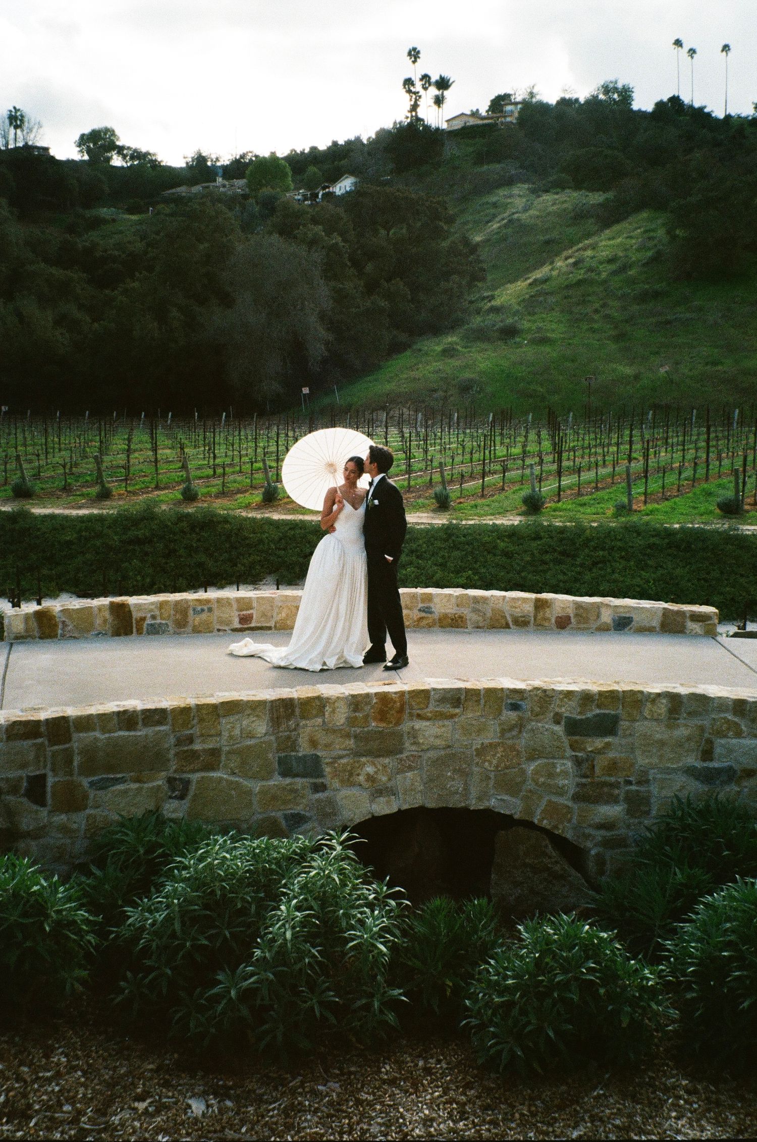 A couple shares a romantic moment with a white parasol on a stone bridge overlooking vineyard rows and green hills.