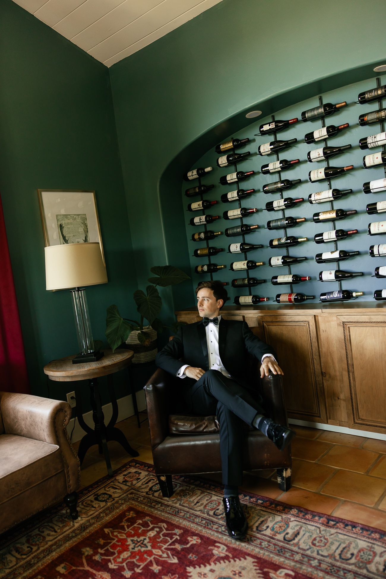 Elegant wine room with dark green walls features a built-in bottle display wall and vintage furniture on a patterned rug.