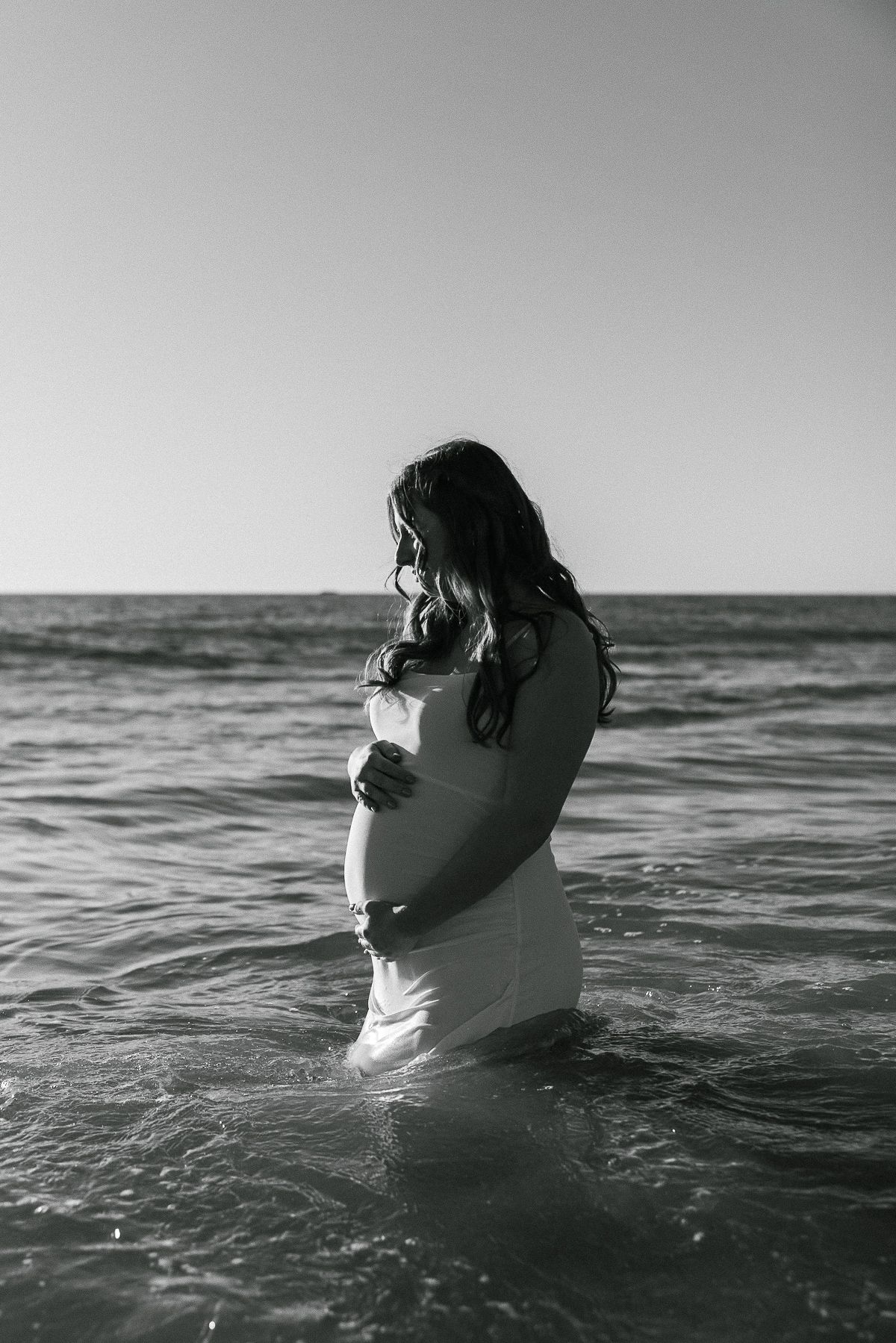 Black and white photo of a pregnant woman standing in ocean waves wearing a flowing dress.