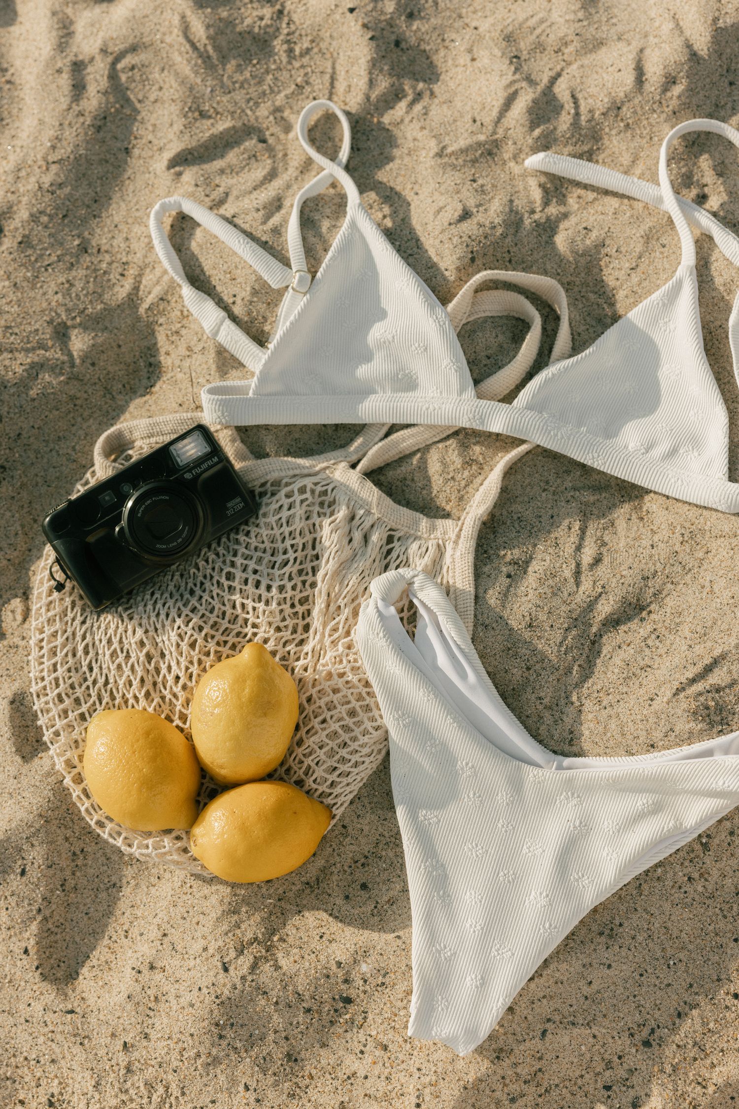 White bikini top and oranges arranged artistically on sandy beach with camera in natural sunlight.