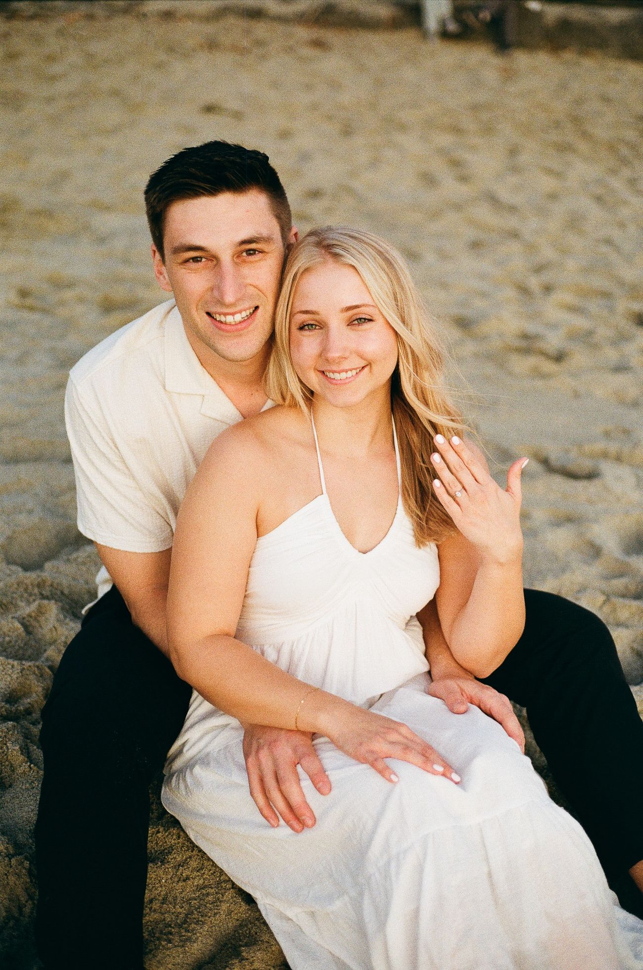 A couple in white outfits smile together while sitting on a sandy beach during a romantic photoshoot.