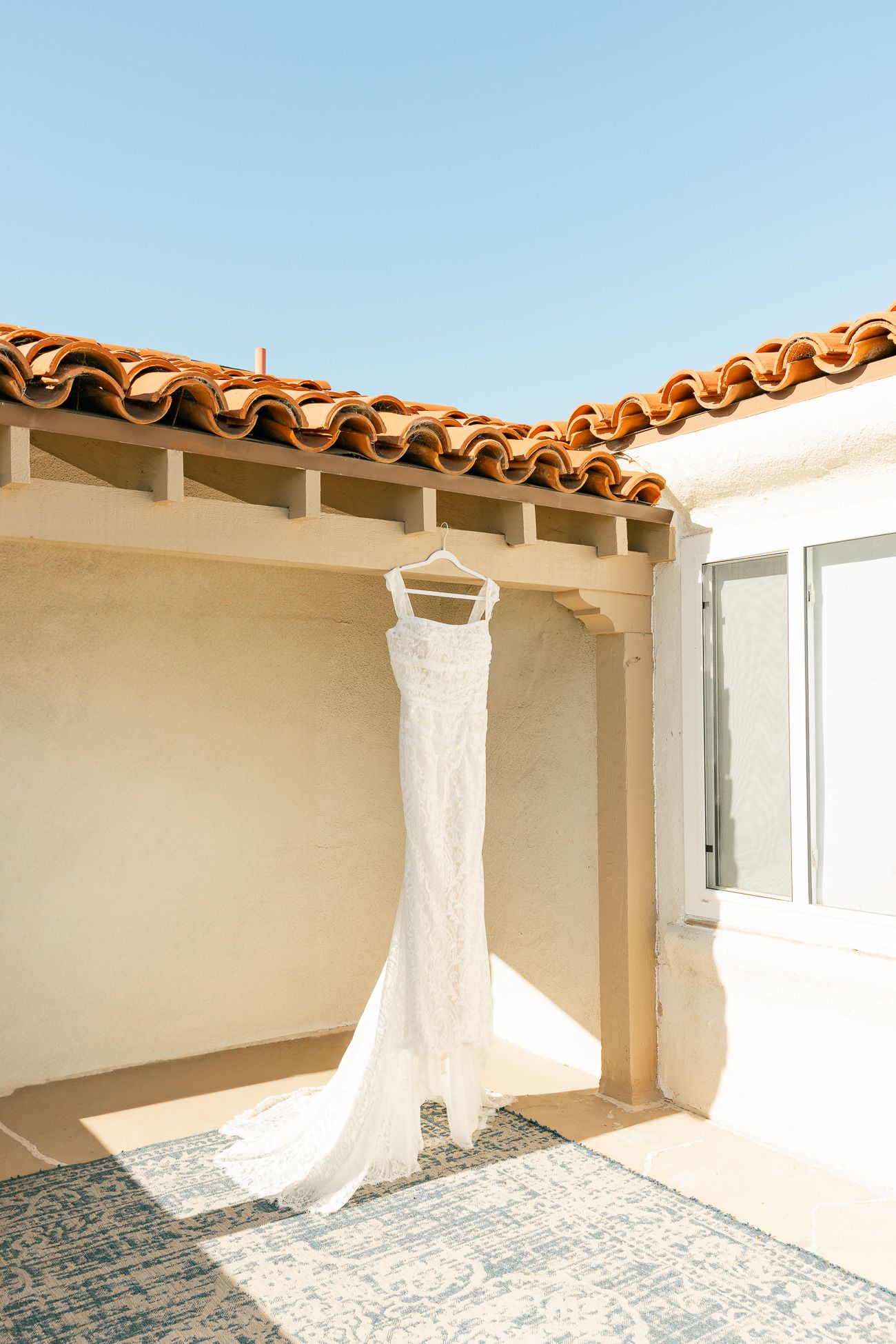 A white lace wedding dress hangs against a cream wall under terracotta roof tiles in Mediterranean sunlight.