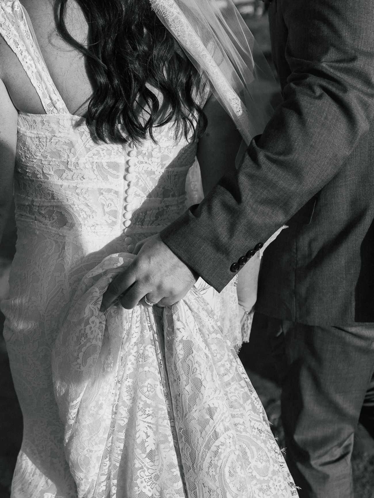A close-up black and white photo showing intricate lace details of a wedding dress and a suit sleeve.