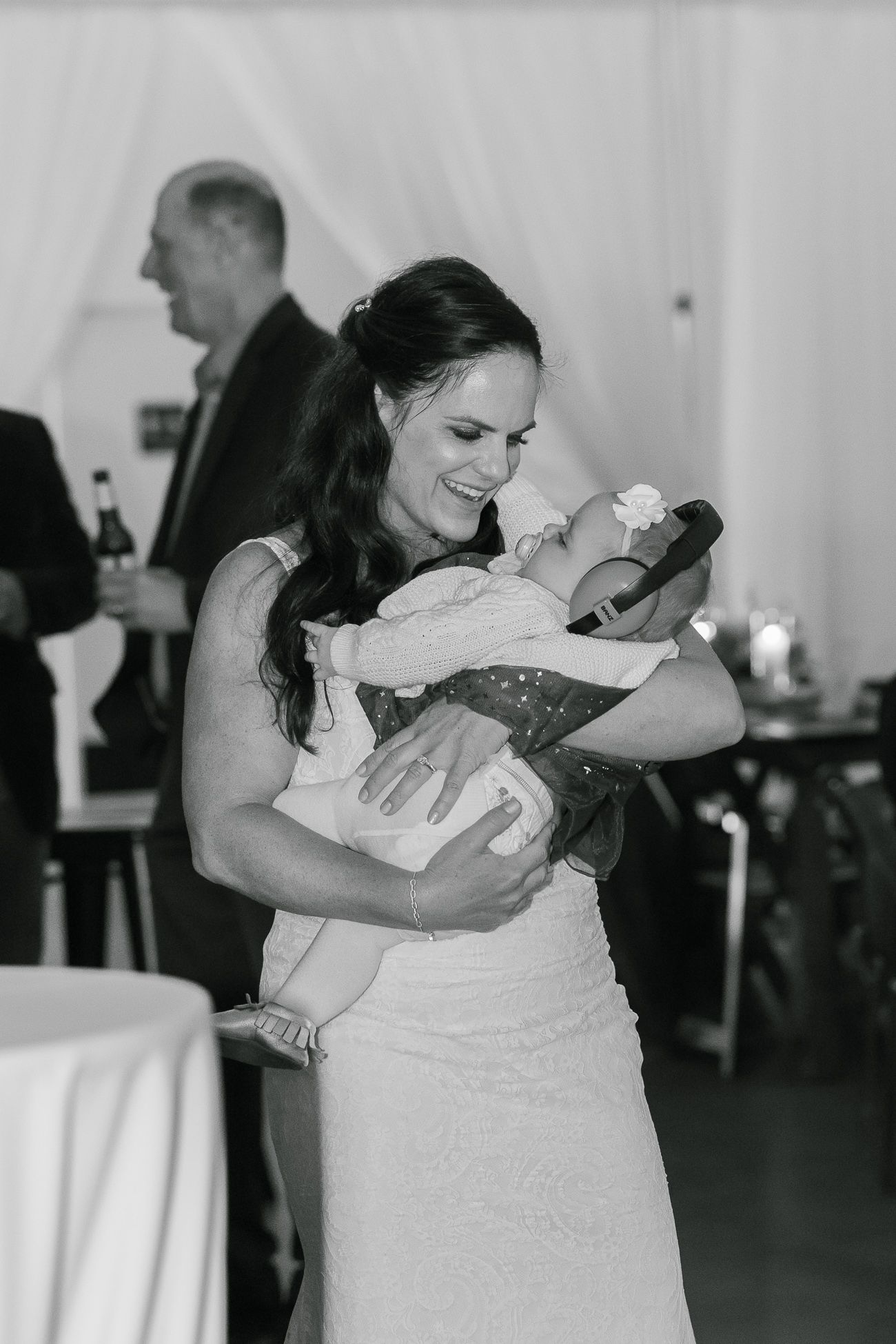 A woman in a white dress holds and gazes lovingly at a baby during a formal event.