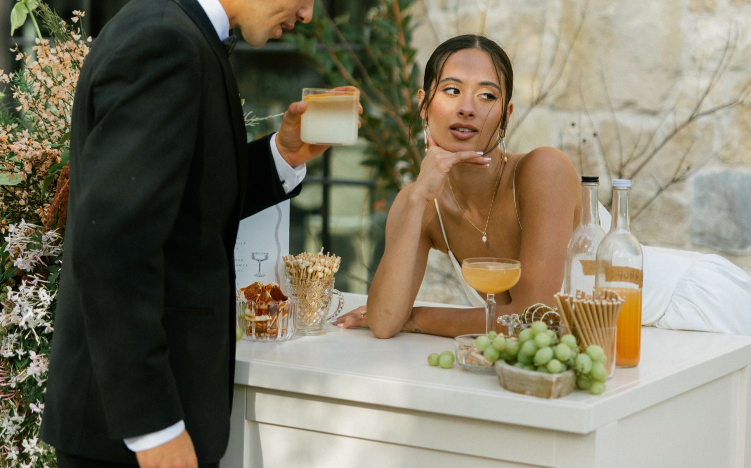 A restaurant scene with drinks and grapes on a white table as a server interacts with a guest dressed in formal attire.