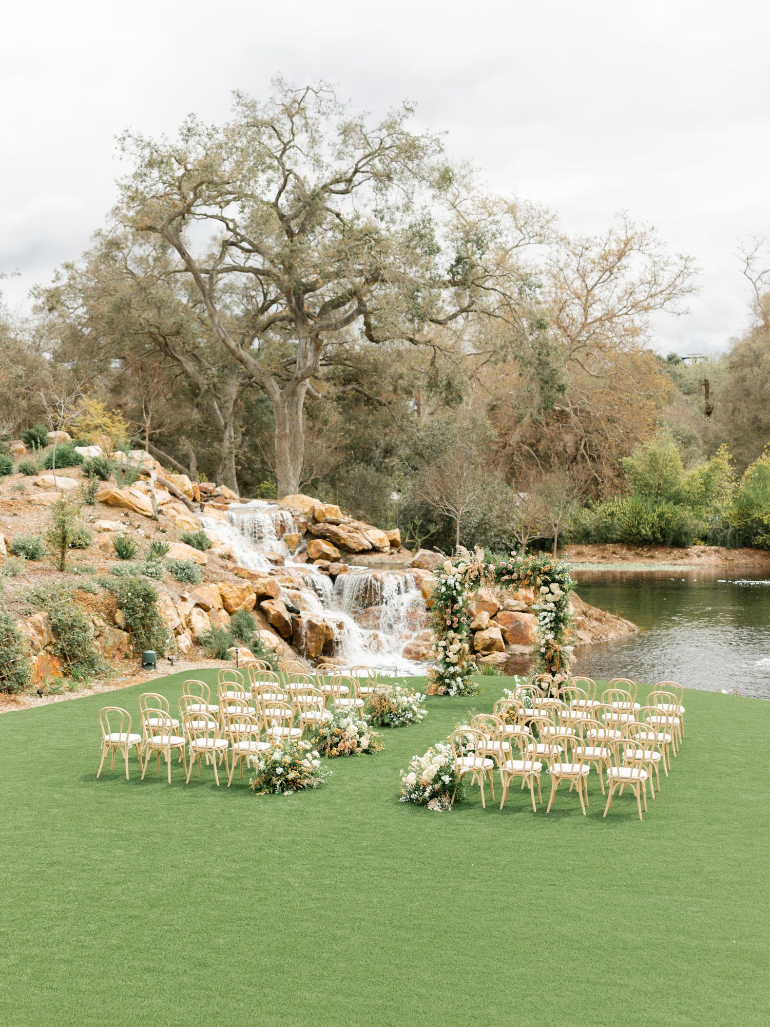 Outdoor wedding ceremony setup with wooden chairs on manicured lawn facing a natural waterfall and trees in the background.
