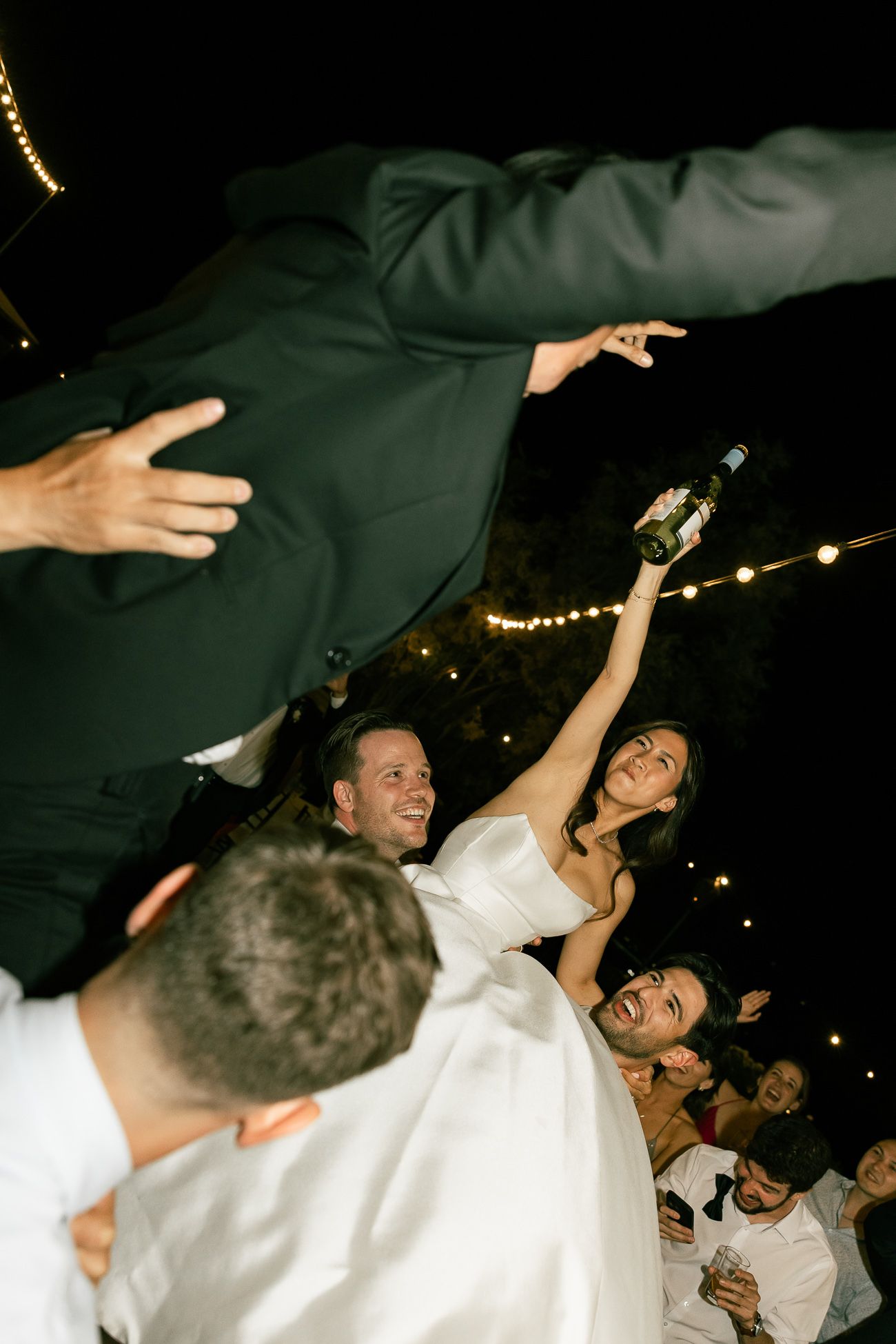 A group of wedding guests dancing and celebrating at a reception with string lights in the background.