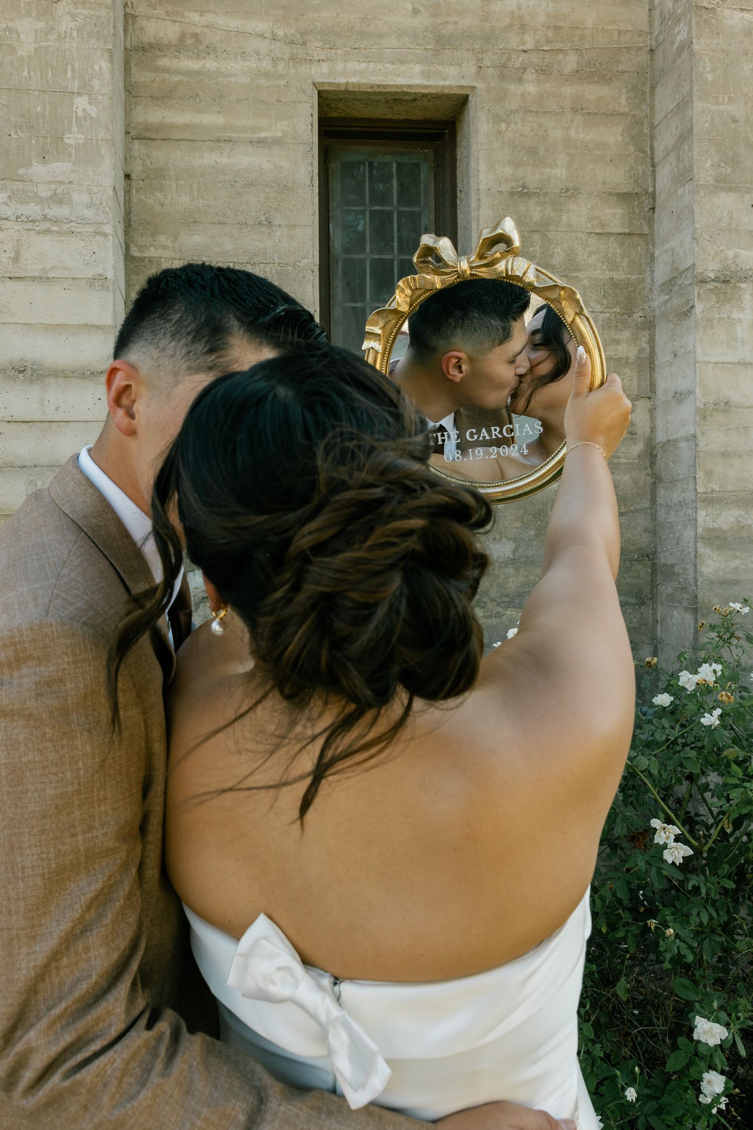 A couple shares an intimate moment on their wedding day holding a vintage mirror outdoors by a stone wall.