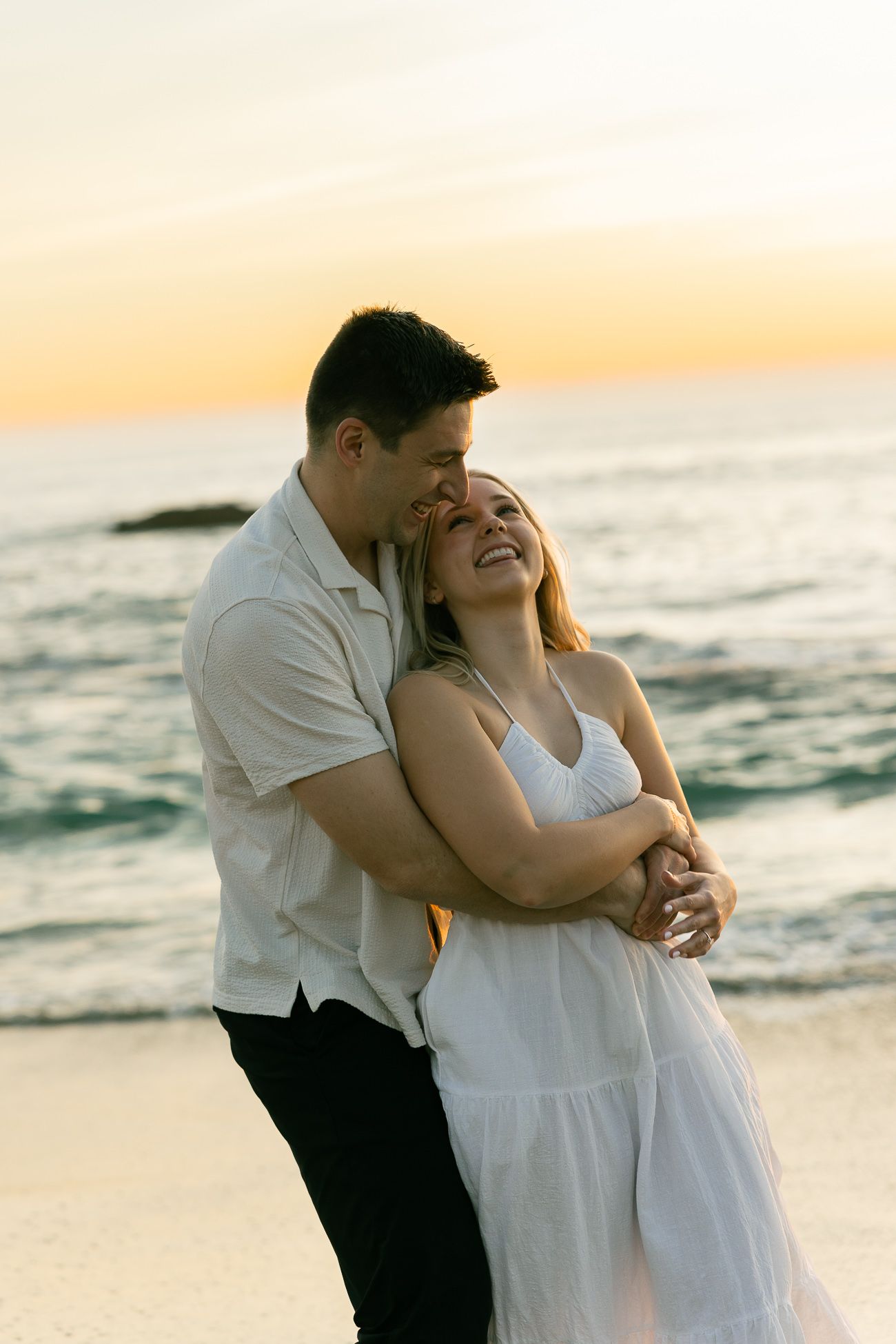 A romantic couple shares a tender moment while laughing together on the beach at sunset.