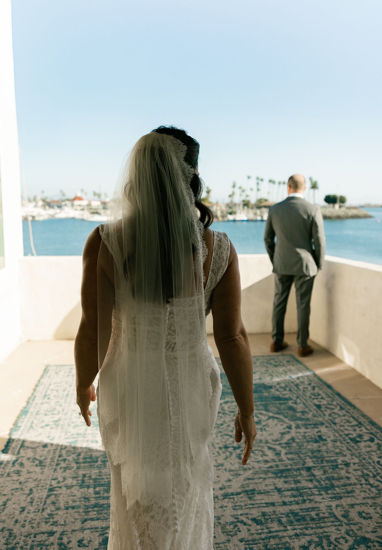 Back view of bride in lace gown approaching groom for first look on oceanfront terrace.