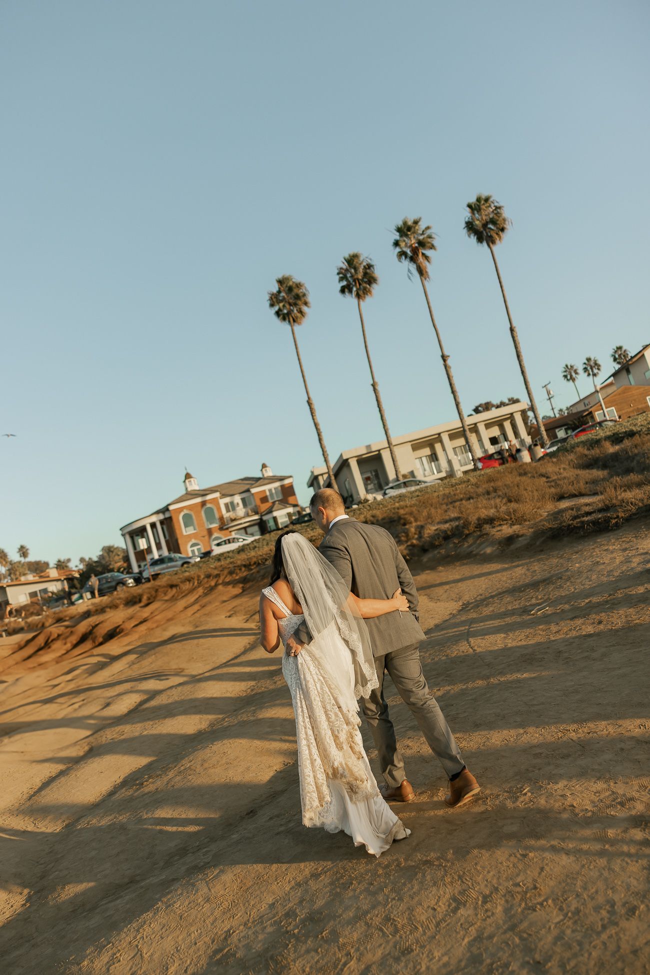 A couple strolls along cliffside path with tall palm trees and coastal buildings in background.