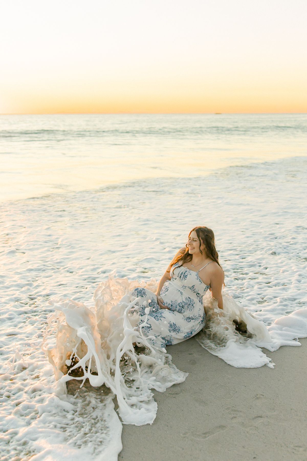 Person in white dress lying on beach sand as ocean waves create dramatic splash effect during sunset.