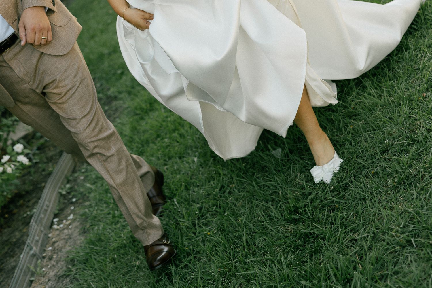 Close-up view of formal shoes and a flowing white wedding dress hem walking on green grass.