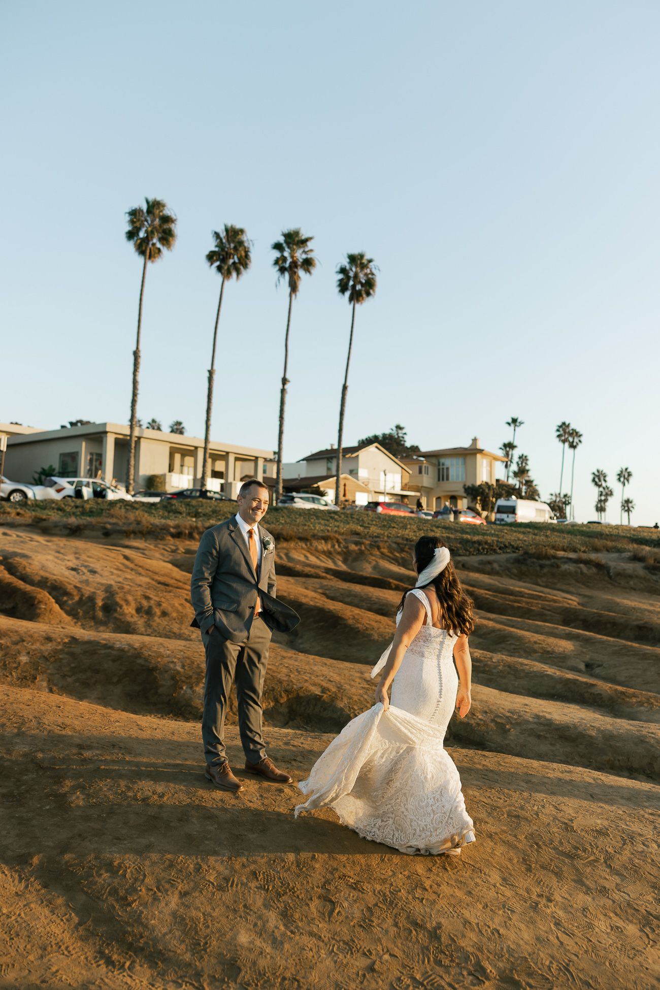A couple in wedding attire stands on a hill with palm trees and coastal homes in background at golden hour.