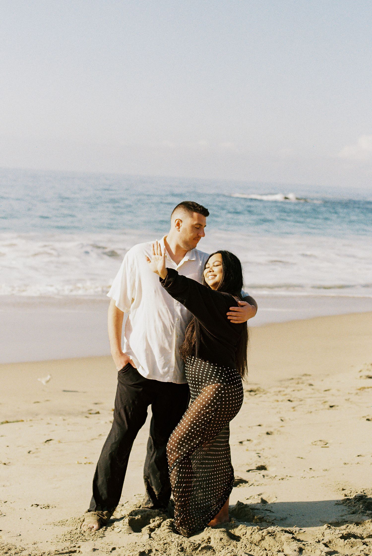 A couple shares a tender moment while sitting together on a sandy beach with ocean waves in the background.