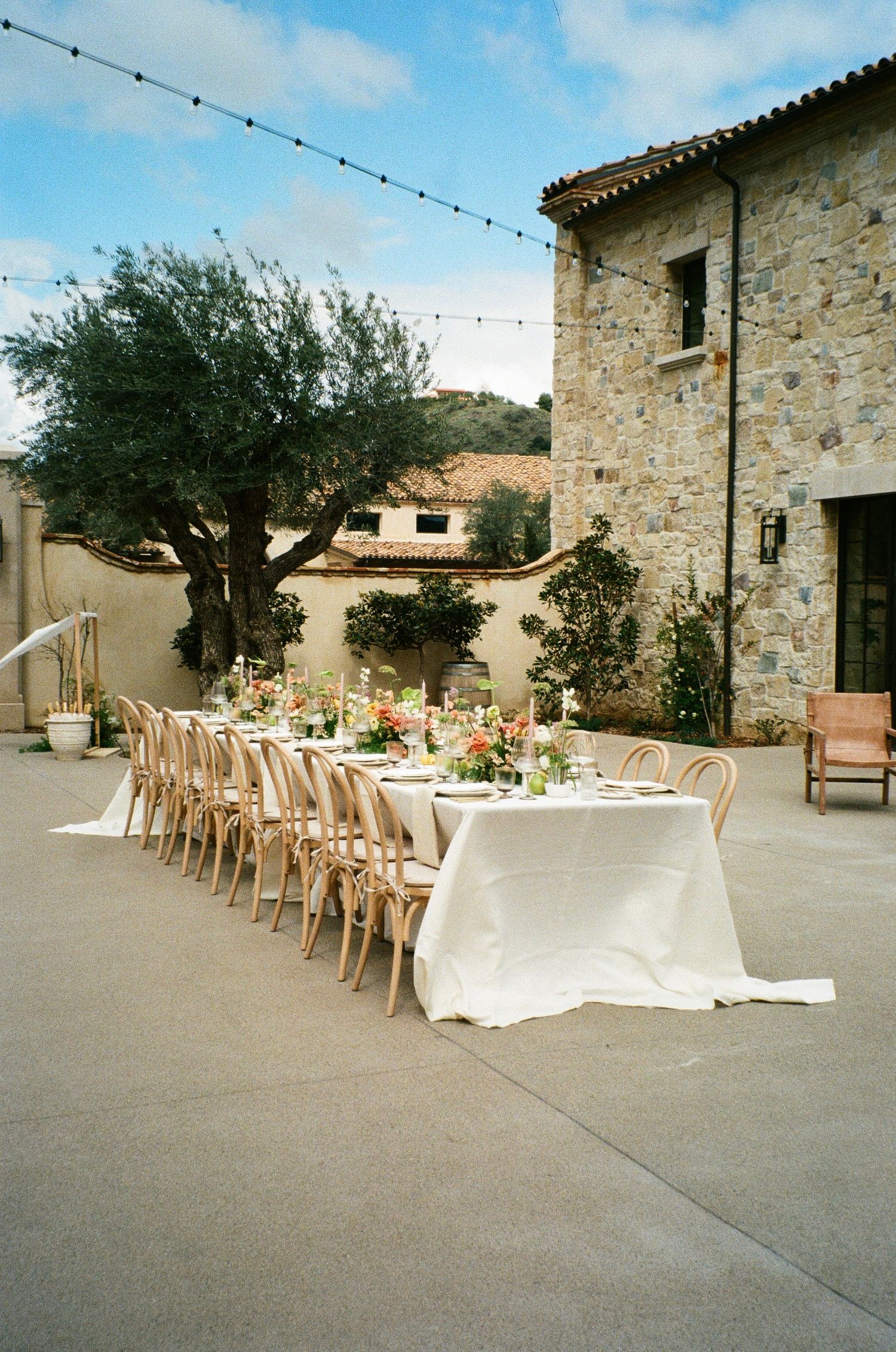 Elegant outdoor dining table with white linens and rattan chairs set against a rustic stone building in Mediterranean style.
