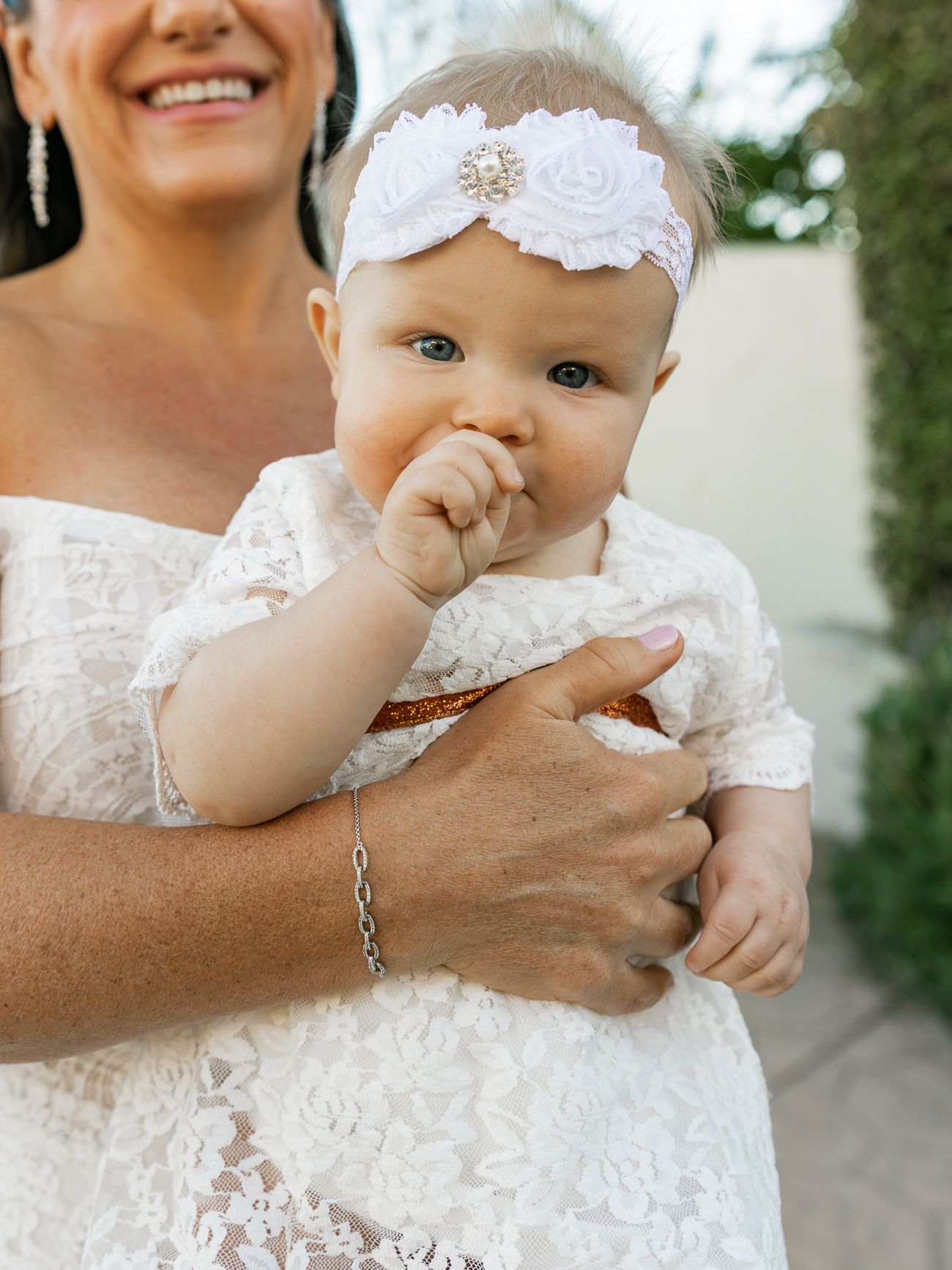 Baby wearing white lace dress and floral headband held during wedding celebration.