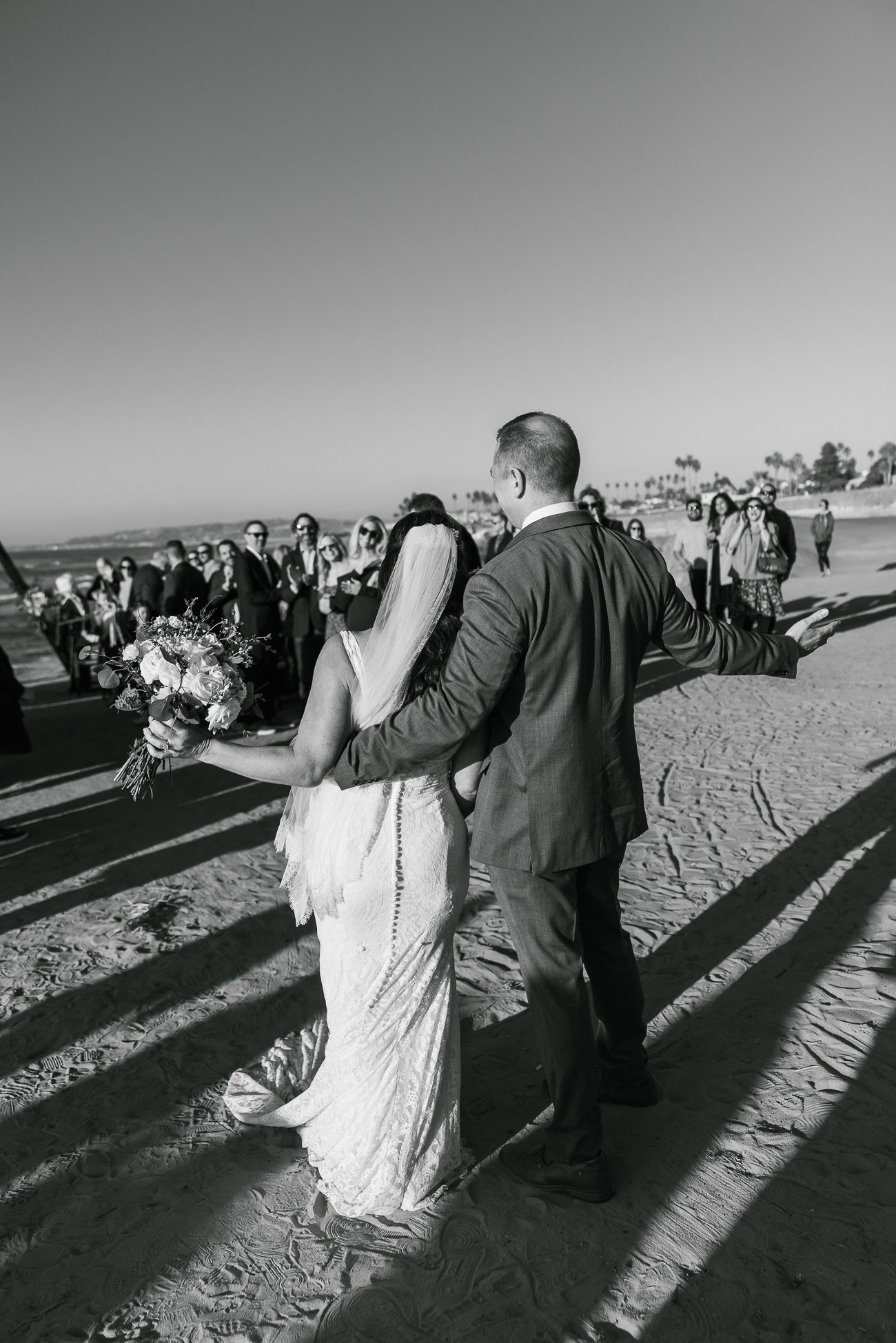 A black and white photo of a couple walking along the beach during their wedding ceremony.